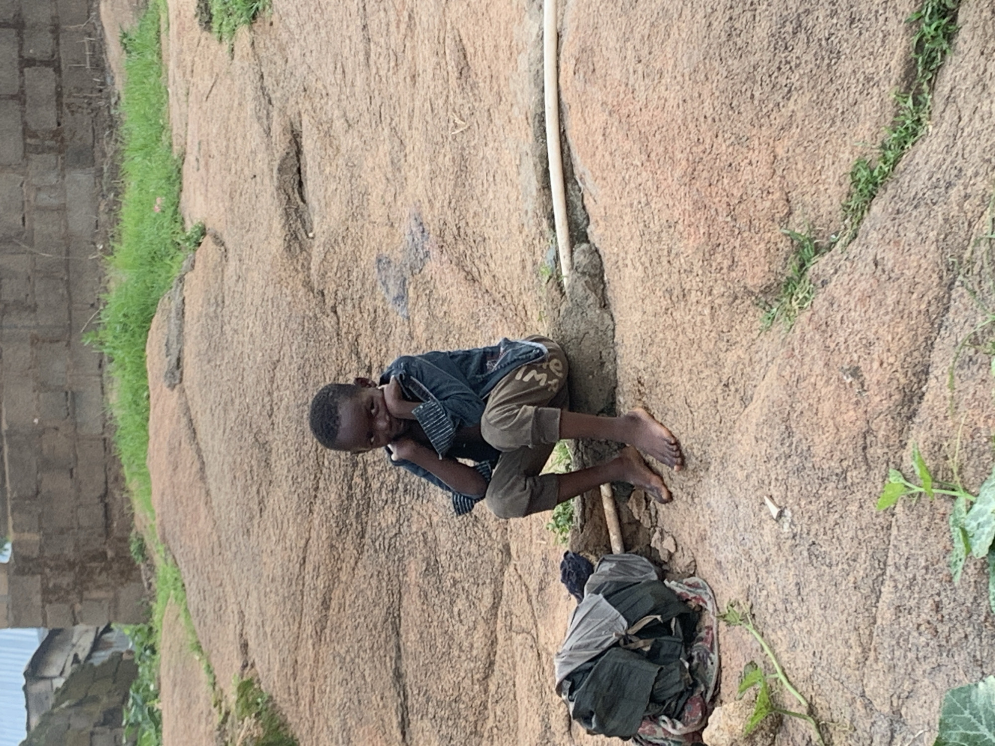 The image depicts a young boy sitting on a rocky surface in what appears to be a rural or semi-urban area. The boy is wearing a dark-colored shirt and light-brown pants, with his elbows resting on his knees and his chin in his hands, suggesting a pensive or somber mood. Beside him, on the left, is a pile of folded clothing. The environment consists of large, textured rocks, patches of dry grass, and a few small green plants. In the background, a brick structure and some makeshift buildings are visible, indicating a human settlement. A white pipe runs horizontally across the middle of the frame. The overall scene conveys a sense of simplicity and perhaps hardship, with the natural elements of the rocks and vegetation contrasting with the man-made elements of the buildings and clothing. The specific location within Jos, Nigeria, cannot be definitively determined from the visual information alone.