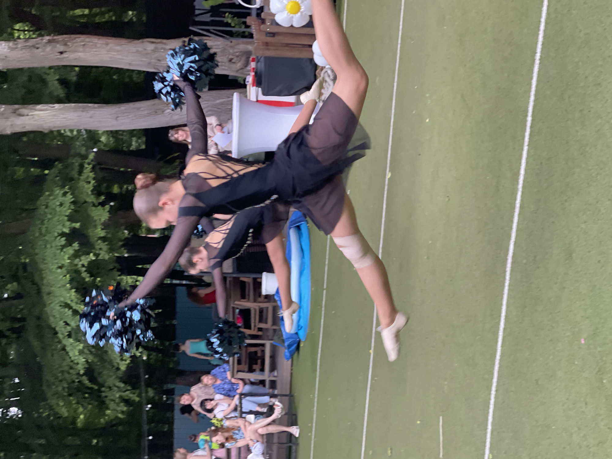Two young women in black performance outfits with sheer skirts are mid-air in a synchronized split leap, holding blue pom-poms. They are on an artificial turf field with white lines, likely a sports field or performance area. In the background, an audience sits on bleachers, with trees and foliage forming a natural backdrop. The scene appears to be outdoors, possibly in a park or an open-air venue in Stavropol, Russia.