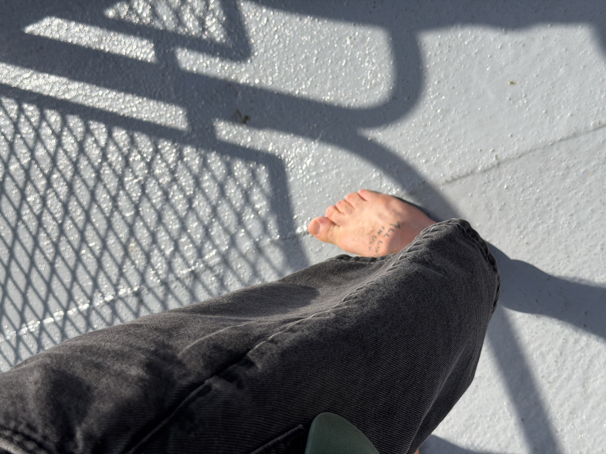 The image presents a first-person perspective of someone sitting on a gray surface, likely a deck or patio. The person is wearing dark gray jeans, and their bare foot is visible. The foot has a tattoo of illegible writing. The deck surface has a textured, possibly non-slip finish. Sunlight casts shadows from a nearby metal mesh structure onto the deck surface. The location, Boston, United States of America, cannot be confirmed from the visual information alone. There are no interactions or actions visible in the picture.
