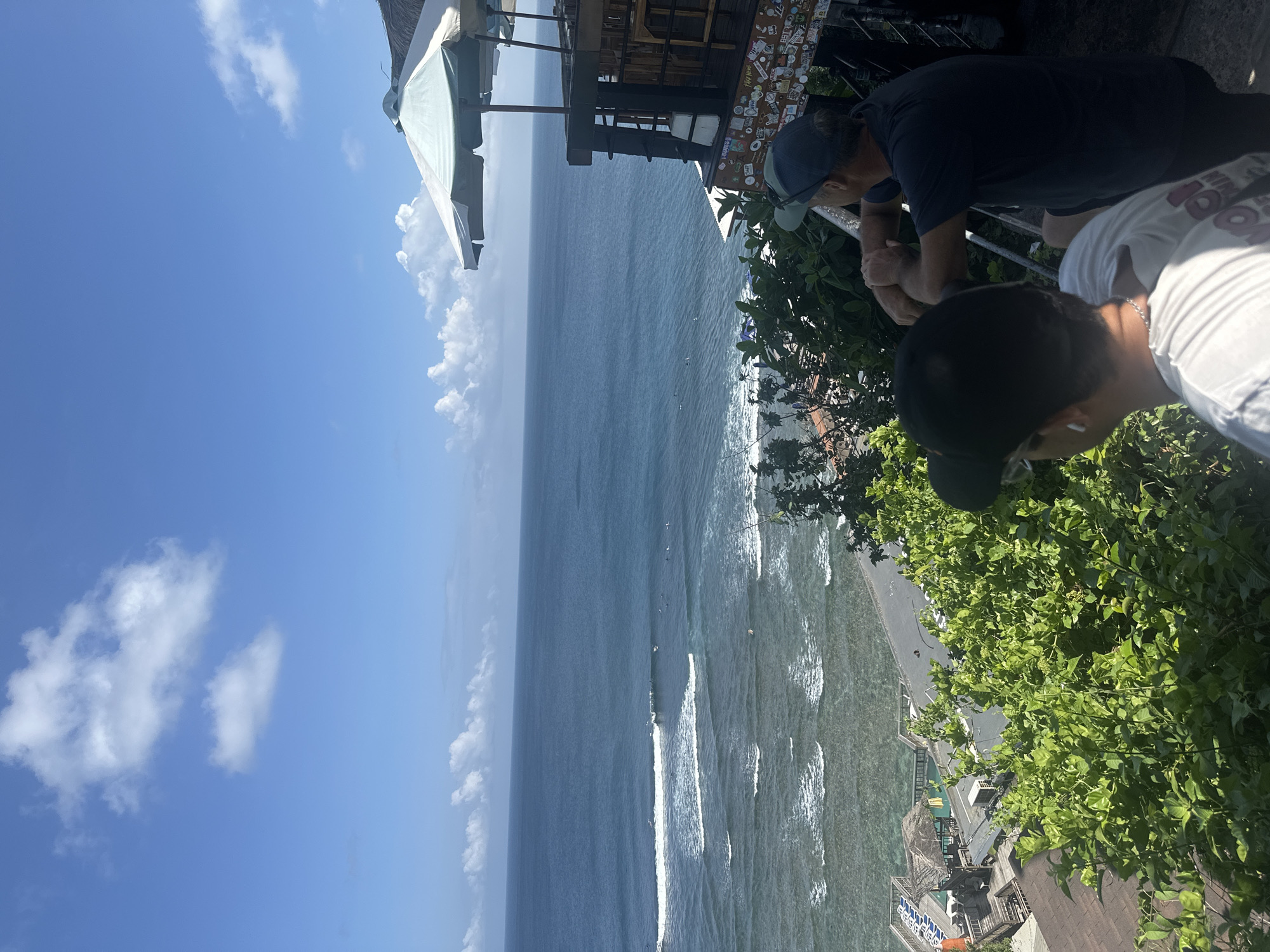 The image is a high-angle view overlooking the ocean from a structure in Kuta Selatan, Indonesia. The ocean spans the majority of the image, with white waves visible near the shoreline. Two people are visible, both leaning over a railing. One person is wearing a dark-colored shirt and a baseball cap. The other person is wearing a light-colored shirt, a cap, and has a chain necklace. Foliage, primarily green bushes, are abundant on the right side of the frame. Part of the structure includes a white sunshade, a balcony and an exterior wall covered with stickers. The sky in the background is blue with scattered white clouds.