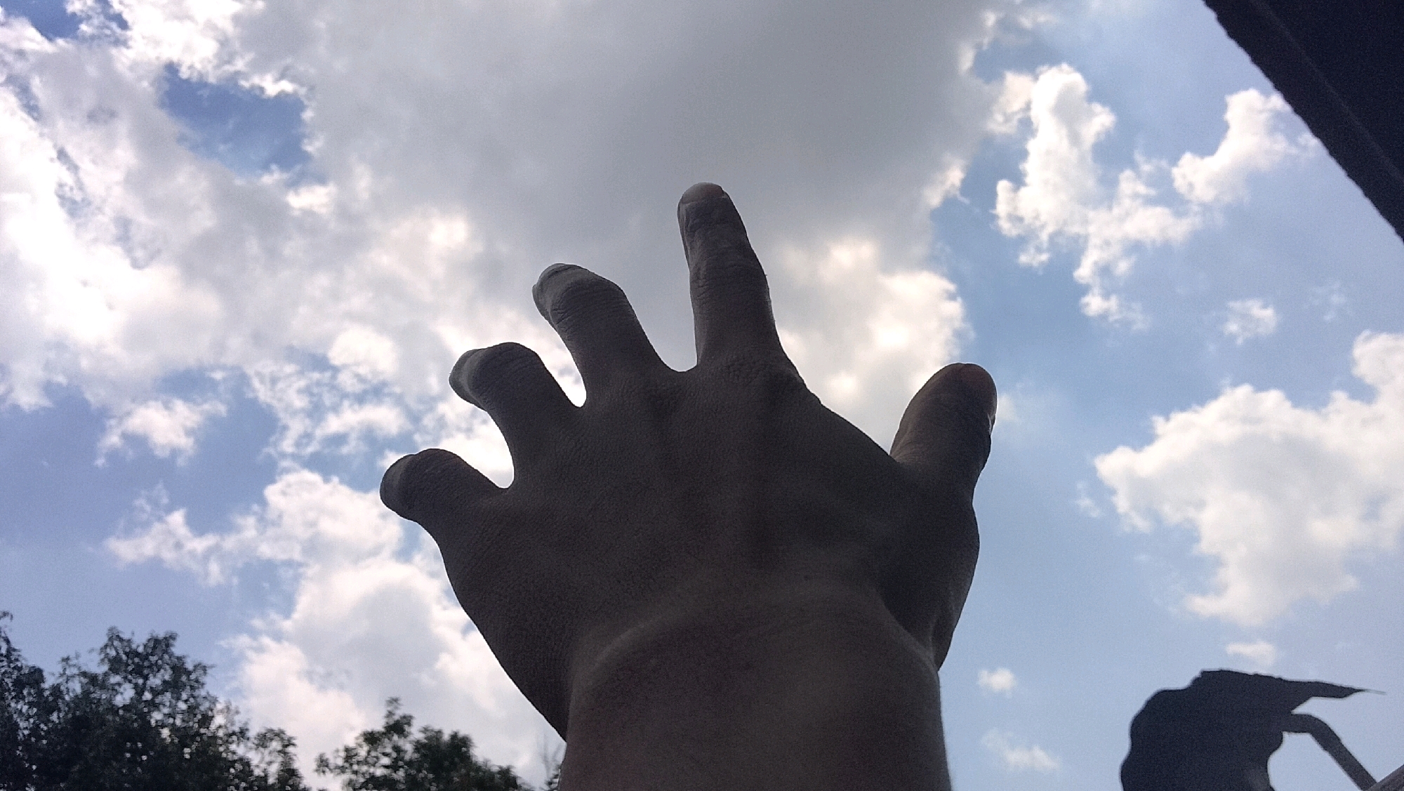 A dark, silhouetted human hand is raised against a bright, cloudy sky. The palm is facing the viewer, and the fingers are spread apart, with the index finger pointing upwards. The hand appears to be in focus, while the sky and the hint of trees at the bottom of the frame are slightly blurred. The lighting suggests a sunny day with scattered clouds. The image composition is from a low-angle perspective, looking up at the hand and sky. There is a dark, irregular shape in the lower right corner, possibly part of a building or another object. The specific location of Bojong Barat, Indonesia, cannot be determined from the visual information in the image.