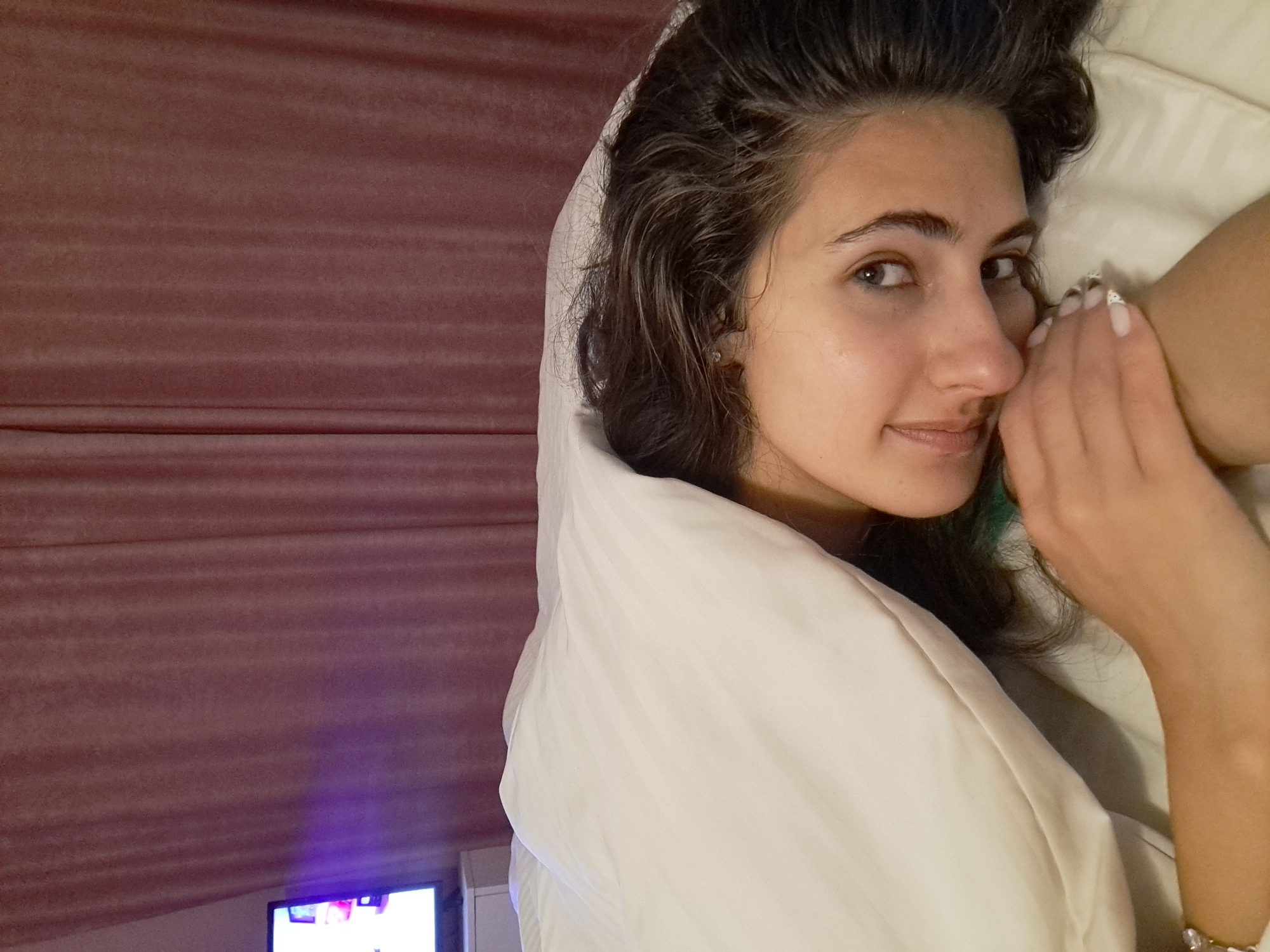 A young woman is lying in bed, partially covered by a white blanket or duvet. She is looking at the camera with a slight smile. Her hair is dark and appears damp, suggesting she may have recently showered. She is wearing a bracelet on her wrist.
Behind her is a pinkish-red textured wall. In the lower left corner, a television is visible, displaying brightly colored images. The background details provide no information about the city of Negovan, Bulgaria.