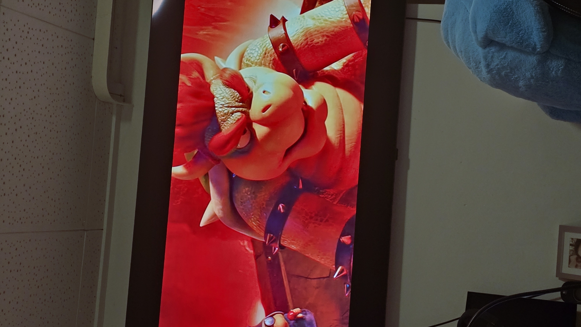 The image depicts a close-up view of a screen displaying a digitally rendered image of Bowser, a character from the Mario franchise. Bowser is rendered in a red hue. The screen is positioned in a room, with a white wall, part of a ceiling with patterned texture, and a blue stuffed animal visible in the background. Parts of a desk and some electronics are visible in the lower corner of the image. There is no indication in the picture of the location being Zirndorf, Germany.