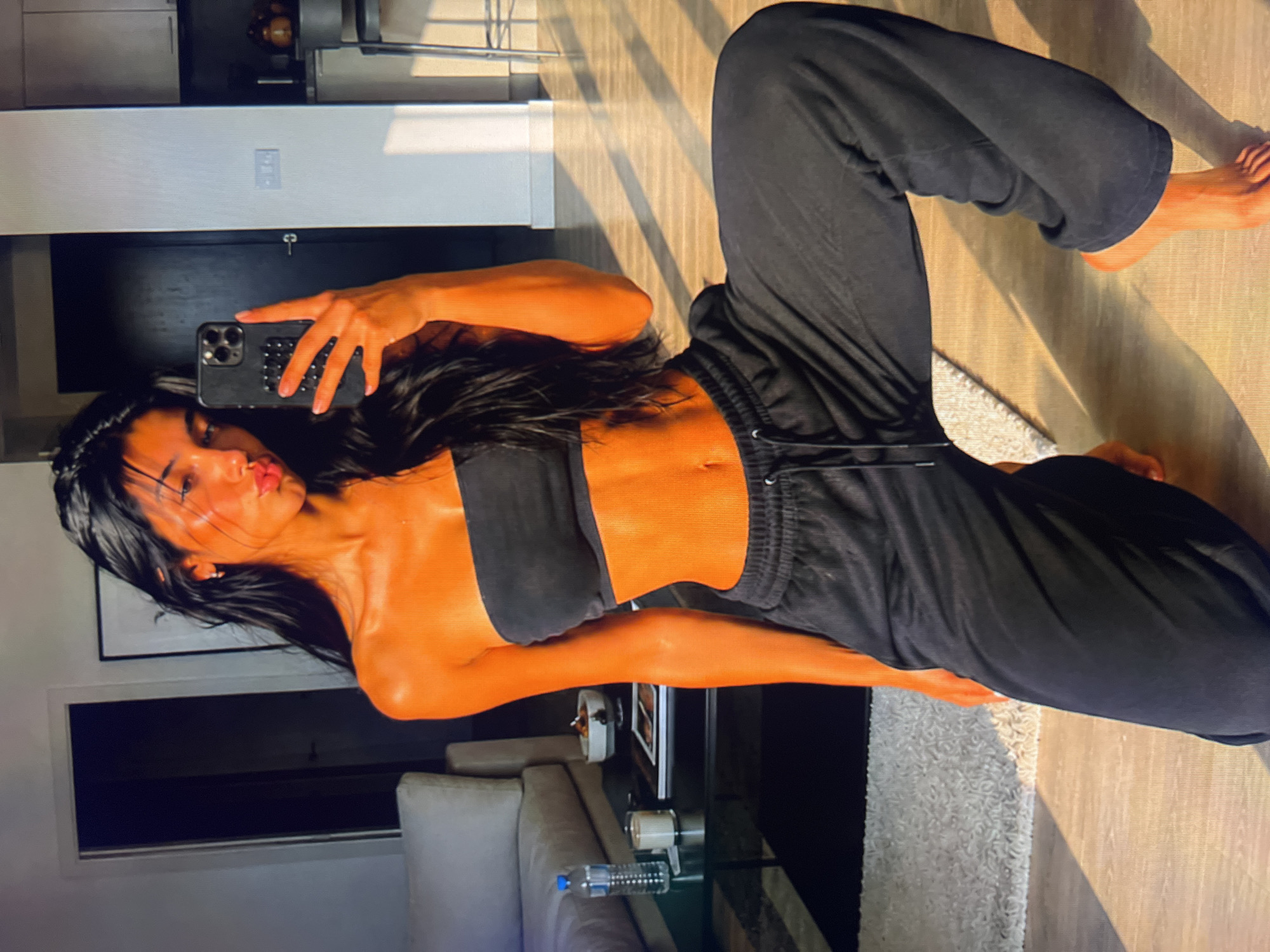 The media file shows a young woman taking a mirror selfie in an indoor setting. She is lying on her side on a wooden floor, wearing black sweatpants and a black bandeau top that exposes her midriff. Her skin has an orange hue, possibly from lighting or a tan. She has long, dark hair and is pursing her lips for the selfie. Her right hand is holding a smartphone, which is positioned to capture the reflection.
The background includes a cream-colored rug and a glimpse of furniture, including a couch and a glass-top table with a bottle of water on it. Sunlight is casting striped shadows across the floor, suggesting it is daytime. The overall mood appears casual and relaxed. The image is captured in portrait orientation, with the subject positioned towards the left side of the frame. There are no visible text elements in the media file.