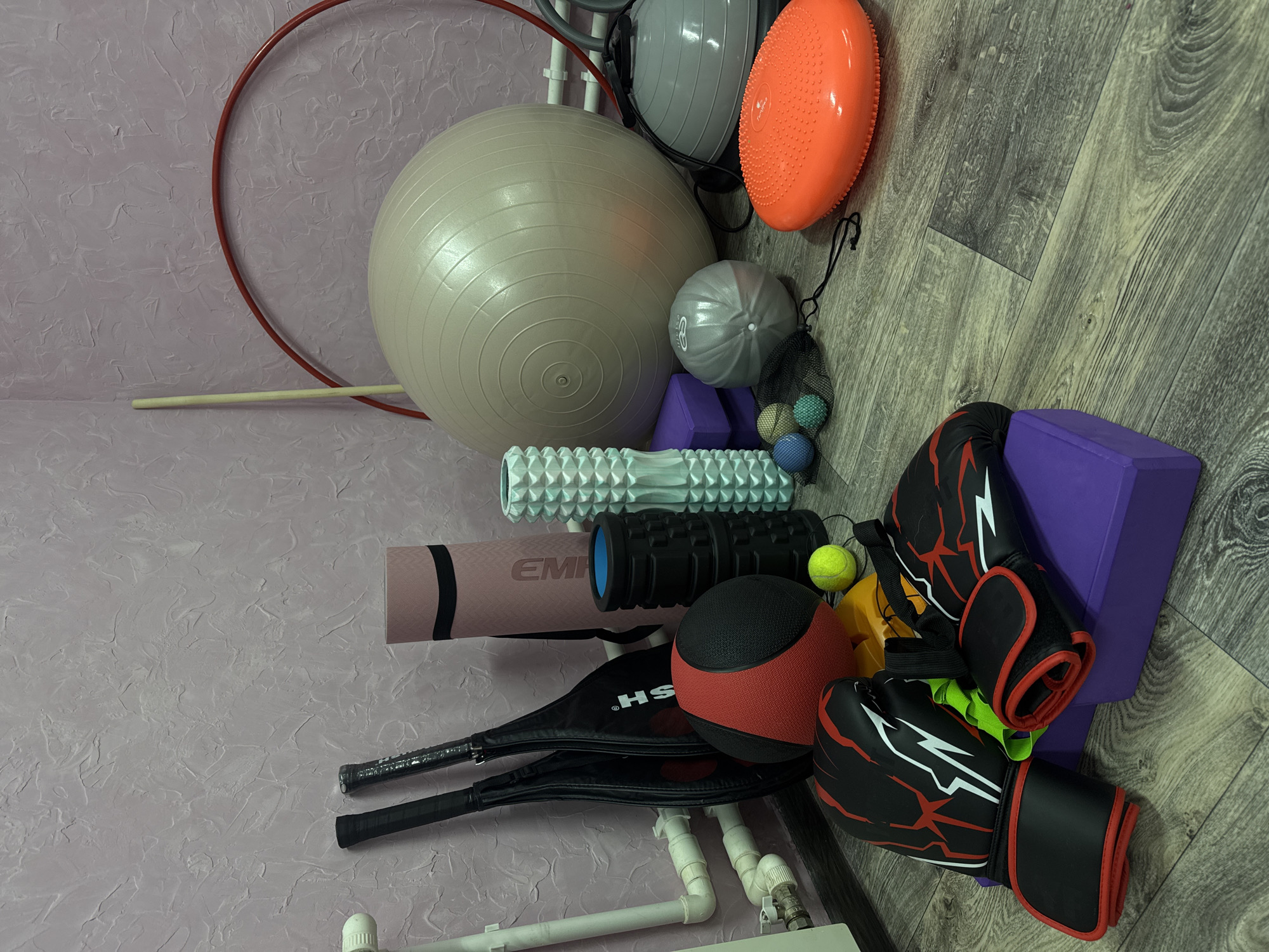 The image shows a collection of fitness-related objects on a wooden floor and against a textured pink wall. The objects include a large inflatable exercise ball, a hula hoop, balance pods, massage rollers, a yoga mat, badminton rackets, a medicine ball, a purple yoga block, and boxing gloves. The location is Прикамский, Russia.