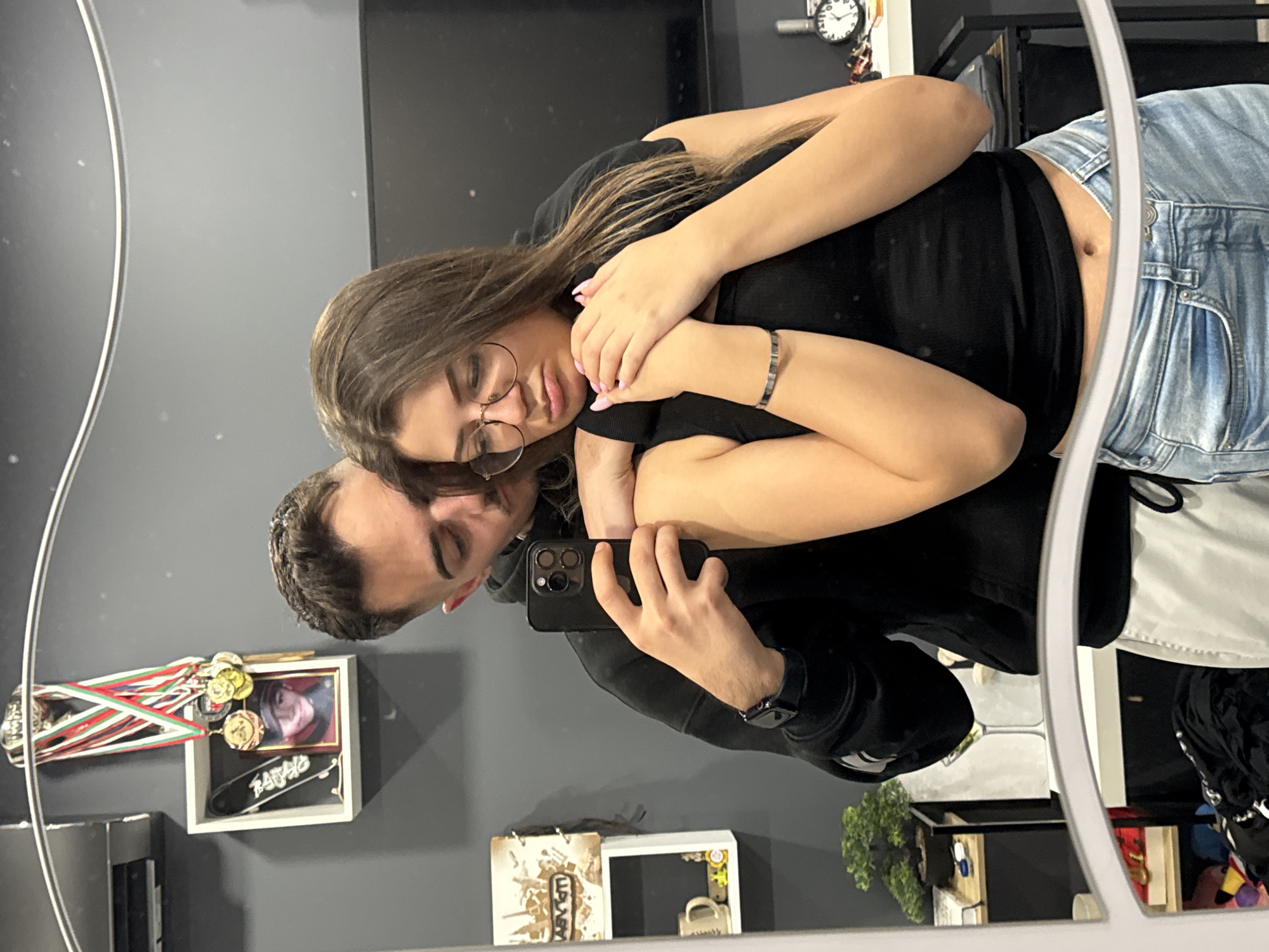 A couple takes a mirror selfie inside a room in Sofia, Bulgaria. The woman wears glasses, a black top, and denim pants. The man, holding the phone, wears a black hoodie and a watch. On the wall behind them are medals, a picture in a frame, and a skateboard. A plant, a book, and a mug are on a shelf below.