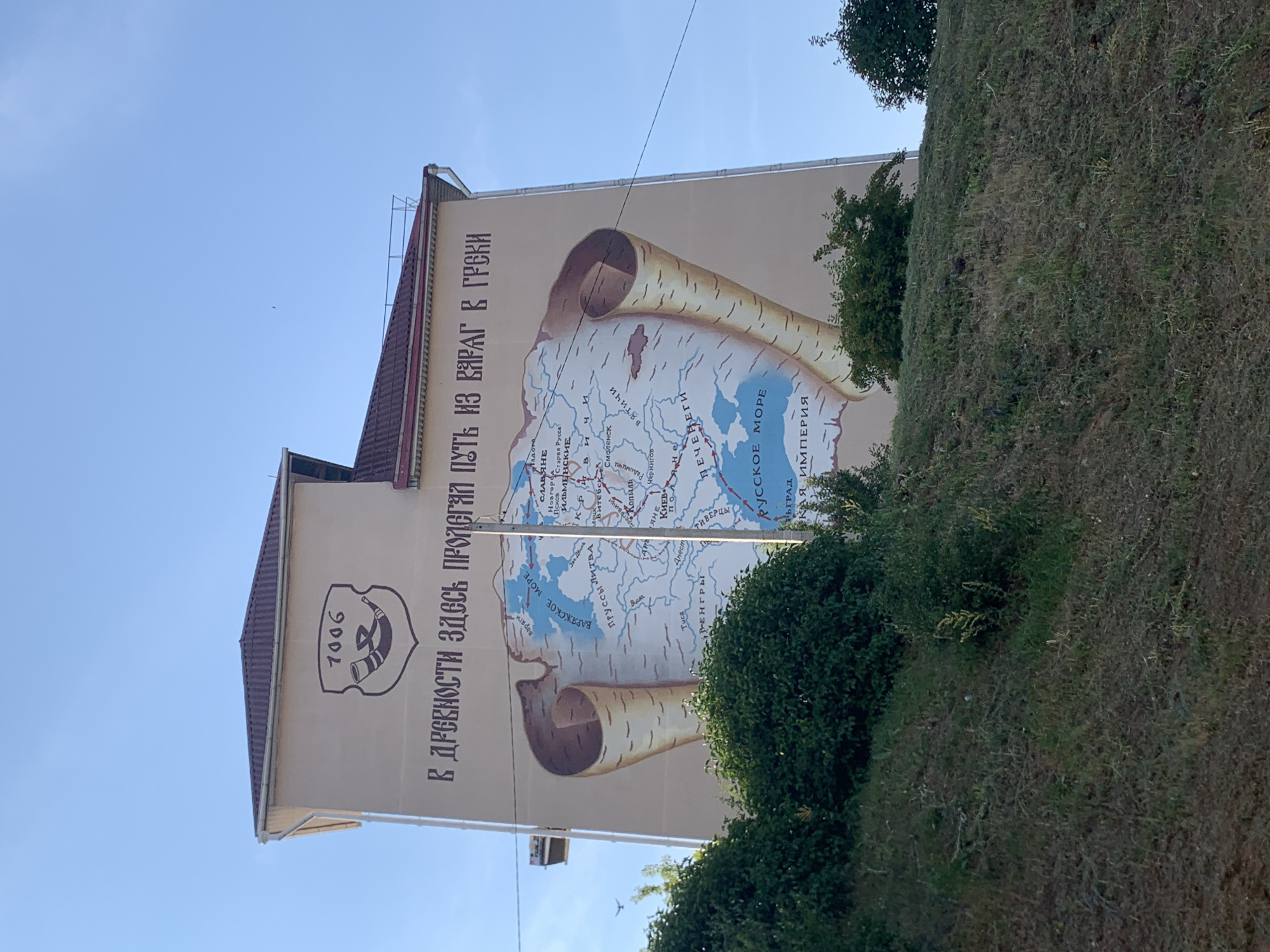 This media file shows a mural painted on the side of a building, depicting a historical map. The mural is oriented vertically, with the top of the building appearing at the bottom of the frame. The mural shows a map with various settlements and bodies of water labeled in Cyrillic script. Two large scrolls, resembling birch bark, frame the map.
The text above the map reads "В ДРЕВНОСТИ ЗДЕСЬ ПРОЛЕГАЛ ПУТЬ ИЗ ВЯРАГ В ГРСКИ," which translates to "In antiquity, the route from the Varangians to the Greeks lay here." This suggests the mural depicts the medieval trade route of the same name.
On the left side of the building, below the mural, is a crest with the number "7006" and what appears to be a horn or trumpet. The overall scene suggests an outdoor setting on a sunny day with a clear blue sky. Green foliage and trees are visible in the foreground and partially obscuring the lower portion of the building. The perspective is from below, looking up at the mural. The image has been rotated from its original orientation for display. The location context is Kapyĺ, Belarus.