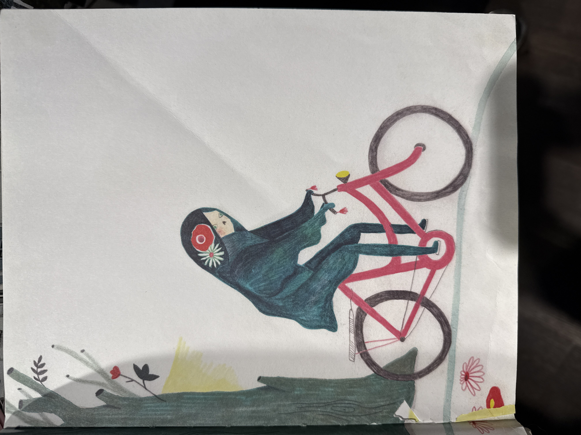 An illustrated image depicts a stylized figure on a bicycle, set against a plain, light-colored background. The main subject is a person, likely a woman or girl, wearing a dark teal or green hooded garment that covers her hair and most of her body, leaving her face visible. She has fair skin and a serene expression. A notable detail on her head covering is a red circular motif with a white daisy-like flower in its center.
She is depicted riding a red bicycle with dark grey tires and a yellow bell or light on the handlebars. The person and bicycle are angled diagonally across the frame, suggesting movement or a dynamic pose, as if suspended in mid-air or falling gently. The person's body is somewhat reclined backward on the bike.
In the bottom section of the image, a large, dark green log or branch lies horizontally, with various small green leaves, reddish flowers, and yellow brush-like elements growing around it. The bottom right corner also features a red mushroom-like object and a white daisy-like flower.
The artwork has a whimsical, illustrative style, possibly created with colored pencils or similar media, evident from the visible texture in the colored areas. There is no visible text in the image. The overall impression is one of imaginative flight or a dream-like scene in a natural, outdoor setting.