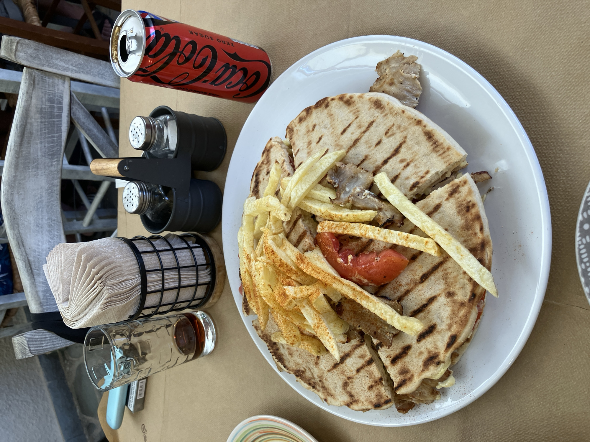 A plate of food sits on a table with a Coca-Cola can and a glass of drink. The food is a gyro with fries and a slice of tomato on top. The plate is white and round. The gyro is wrapped in pita bread that has been grilled. The fries are golden brown and sprinkled with paprika. The Coca-Cola can is red and black and says "Coca-Cola Zero Sugar" on it. The glass is clear and has a dark liquid in it. The table is covered with a brown paper tablecloth. The background is blurry but appears to be an outdoor seating area at a restaurant. A wire napkin holder with napkins, and salt and pepper shakers are also on the table. The scene is set in Agia Marina, Greece.