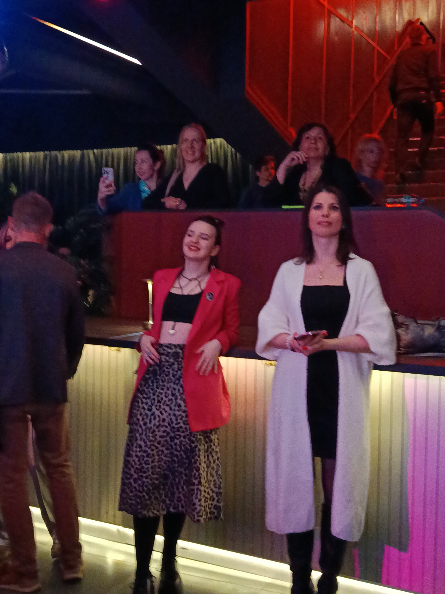 The image depicts a group of people in what appears to be an indoor venue in Moscow, Russia. In the foreground, two women are standing near a bar or counter. The woman on the left is wearing a black crop top, a red blazer, and a leopard print skirt. The woman on the right is wearing a black dress and a long white cardigan. She is holding a smartphone.
Behind them, there is a raised area with a red wall. Several people are seated there, including women taking pictures with their phones. A man is ascending stairs with red railings in the background. A man in a suit can be seen to the left. The venue is dimly lit, with a mix of blue, red, and pink lighting.