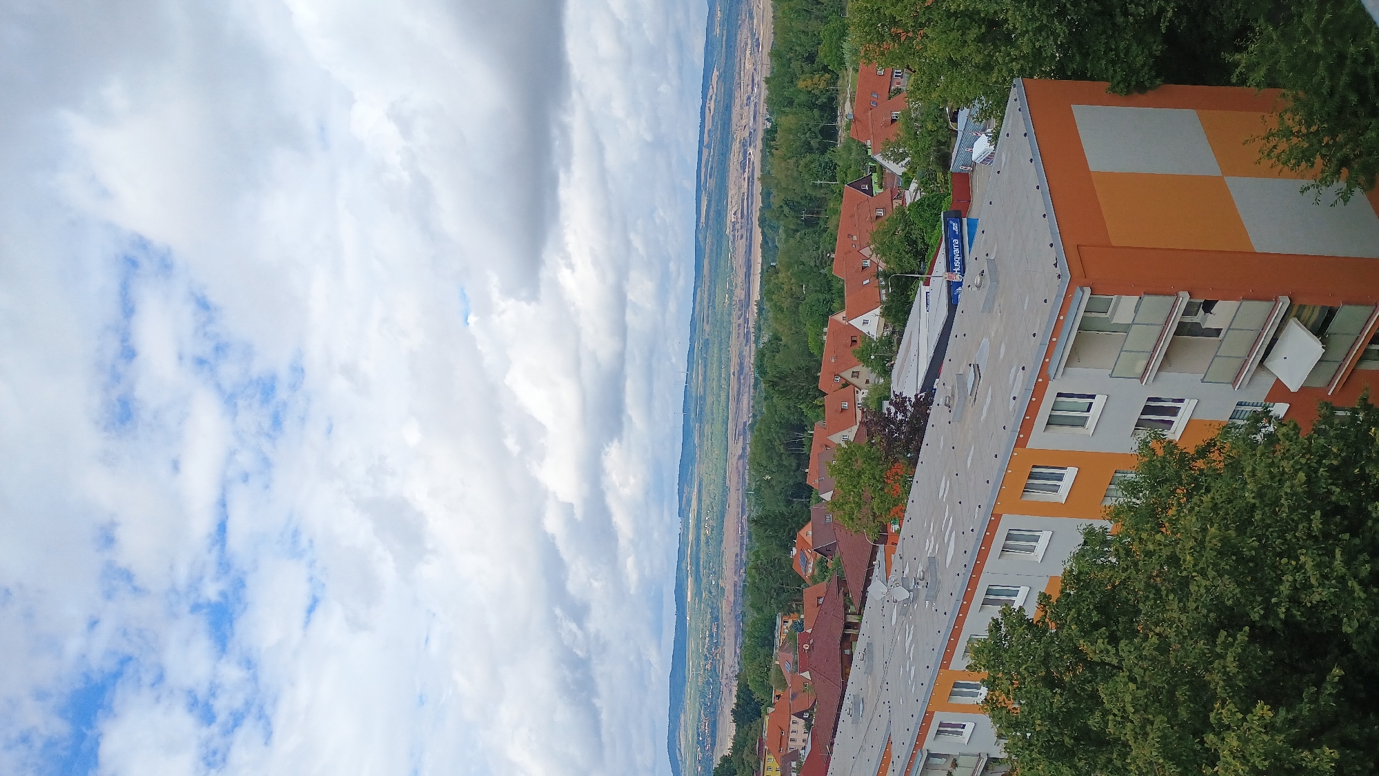 The image captures an elevated view of a residential area in Sokolov, Czechia, with a prominent apartment building in the foreground. The building features an orange and gray facade, with multiple floors of apartments, balconies, and windows. A large green tree partially obscures the left side of the building. Beyond the apartment building, a neighborhood of smaller houses with red roofs is visible, surrounded by lush green trees. In the background, the landscape transitions to a more arid, possibly industrial or mining area, with large exposed earth surfaces and distant hills. The sky above is a mix of blue patches and heavy, dramatic clouds, suggesting overcast or changing weather conditions. A large, open-pit mine or excavation site is clearly visible in the mid-distance, with evidence of industrial activity. Wind turbines are discernible on the horizon, indicating the presence of renewable energy infrastructure. The scene is static, with no visible human activity or interaction.