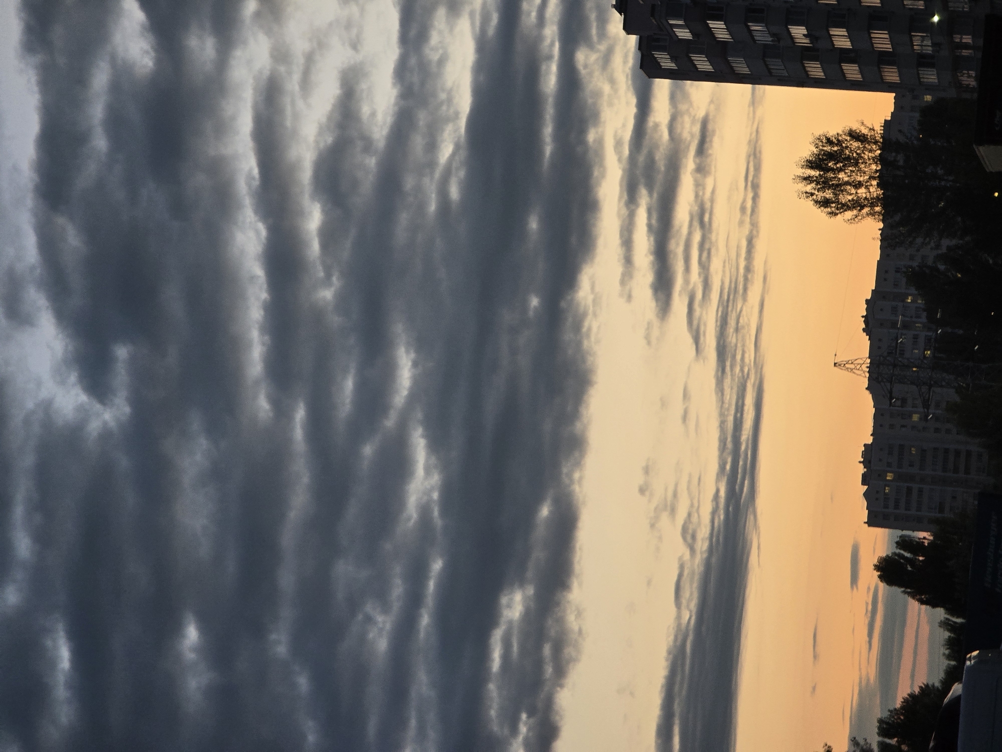 The image captures a twilight sky over Sofiivska Borshchahivka, Ukraine. Dark, layered clouds dominate the upper portion of the frame, with bands of lighter gray and some illuminated edges suggesting the setting sun. Below the clouds, a gradient of warm orange and pale yellow hues fills the sky, transitioning to a deeper, cooler tone near the horizon.
Silhouetted against the vibrant sky are elements of an urban landscape. A tall, multi-story apartment building with numerous windows is prominently featured on the right side of the image. To the left and in the center, the tops of other residential buildings and trees are visible, their forms obscured by the dimming light. A few vertical structures, possibly power lines or cranes, are also faintly discernible. In the foreground, the dark silhouettes of vehicles and what appears to be a dark sign with illegible text can be seen. The overall scene depicts a calm, end-of-day atmosphere in a residential area.