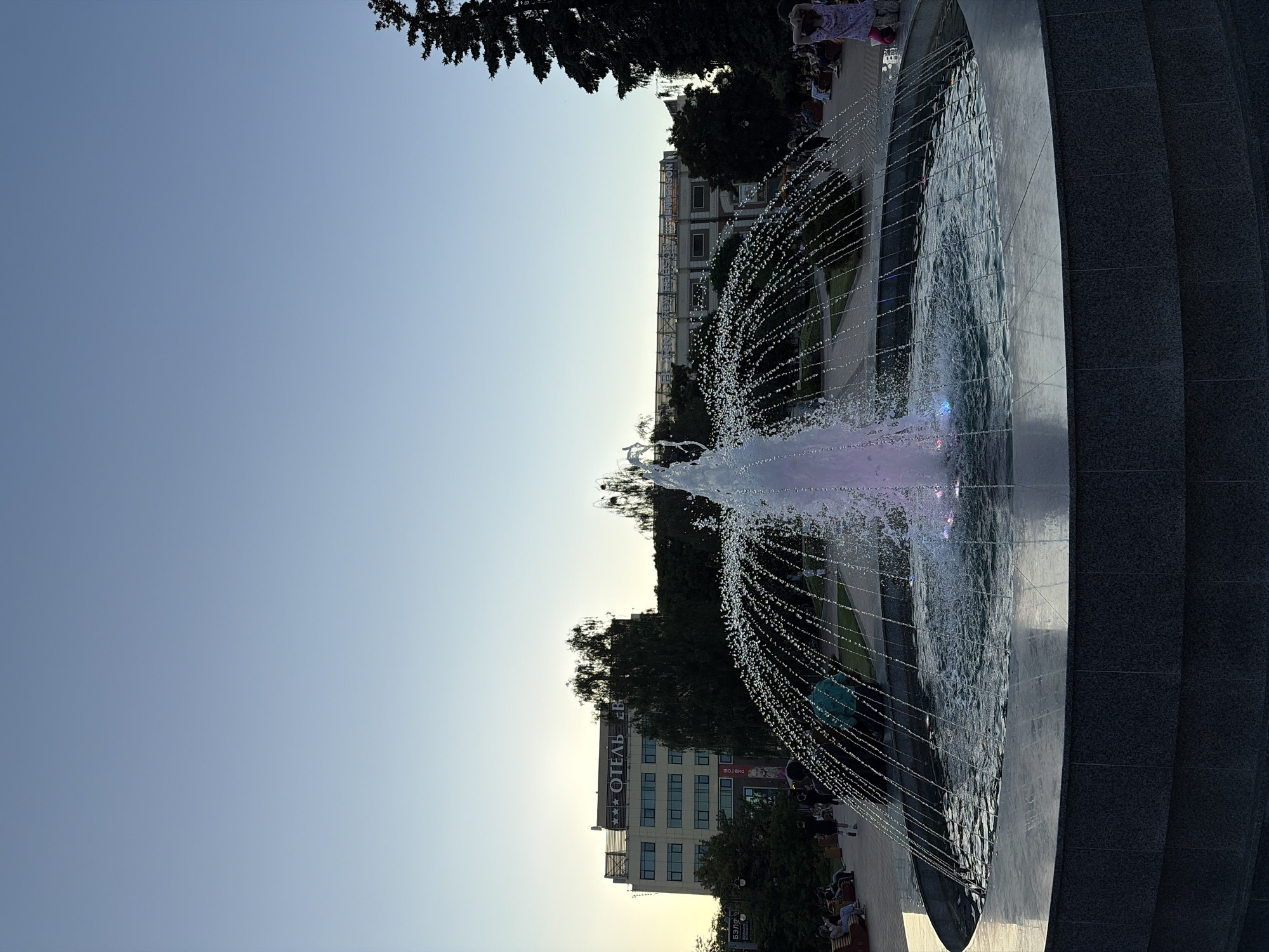 An outdoor, wide-angle shot captures a fountain in the foreground, with a building and trees in the background. The fountain is illuminated with purple and blue lights at its base, and water arcs outward in a circular pattern, with fine droplets catching the light. The sky above is a clear, light blue, indicating it is likely dusk or dawn.
In the background, a multi-story building with a sign that reads "ОТЕЛЬ" (Hotel) is visible. Trees surround the building and extend into the distance. A few people are scattered throughout the scene, some sitting on benches and others walking. On the right side of the frame, a person in a light-colored dress and pink pants stands with their hands behind their head. On the left, several individuals are seated on park benches, observing the fountain. The environment appears to be a public park or square.