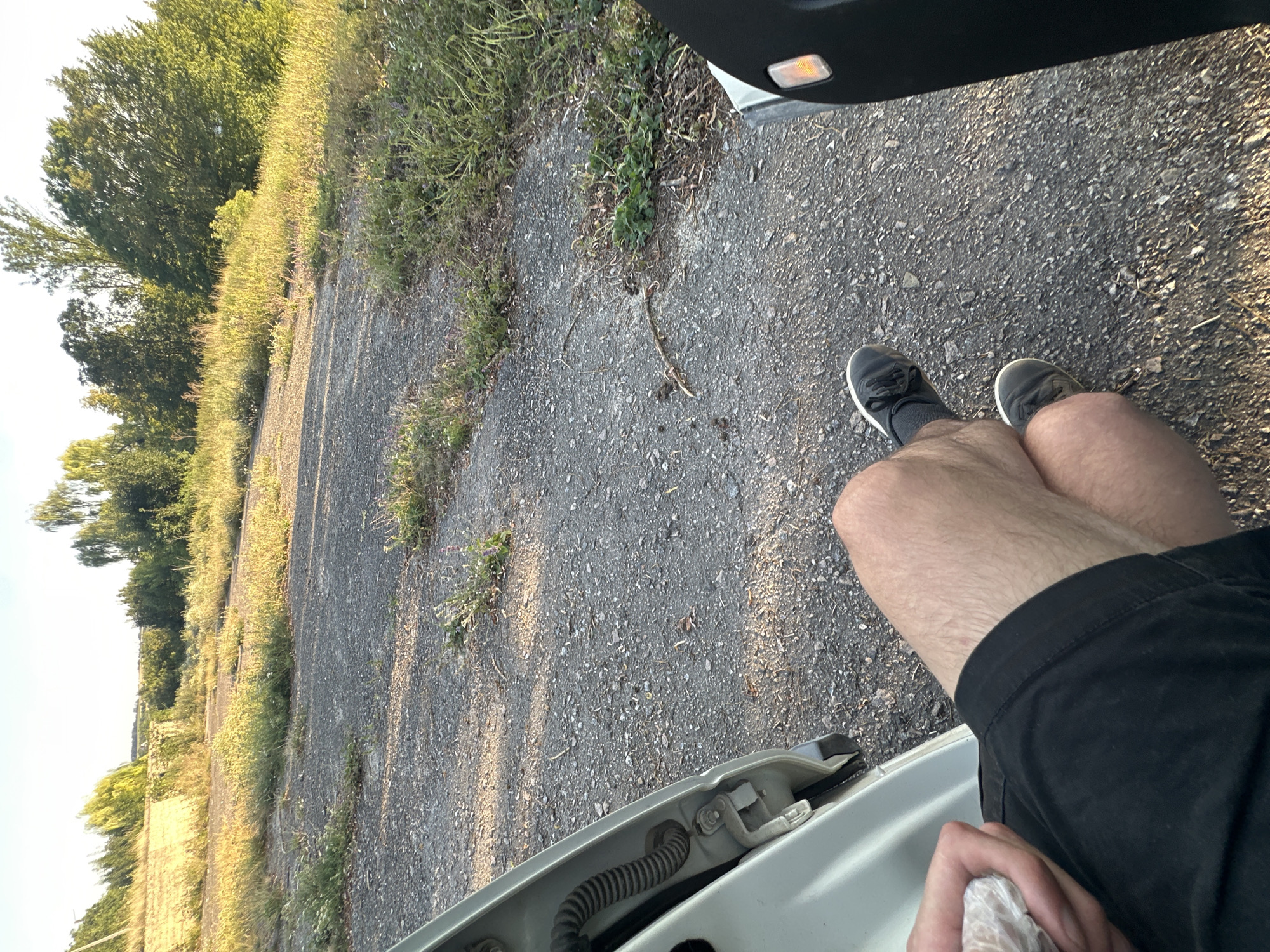 The image shows a person sitting with their legs out of the open door of a car, on a paved surface with sparse vegetation. The background consists of trees and a clear sky. The person is wearing black shorts and dark sneakers with white soles. They are holding a white object in their left hand. The location is identified as Makiv, Ukraine.