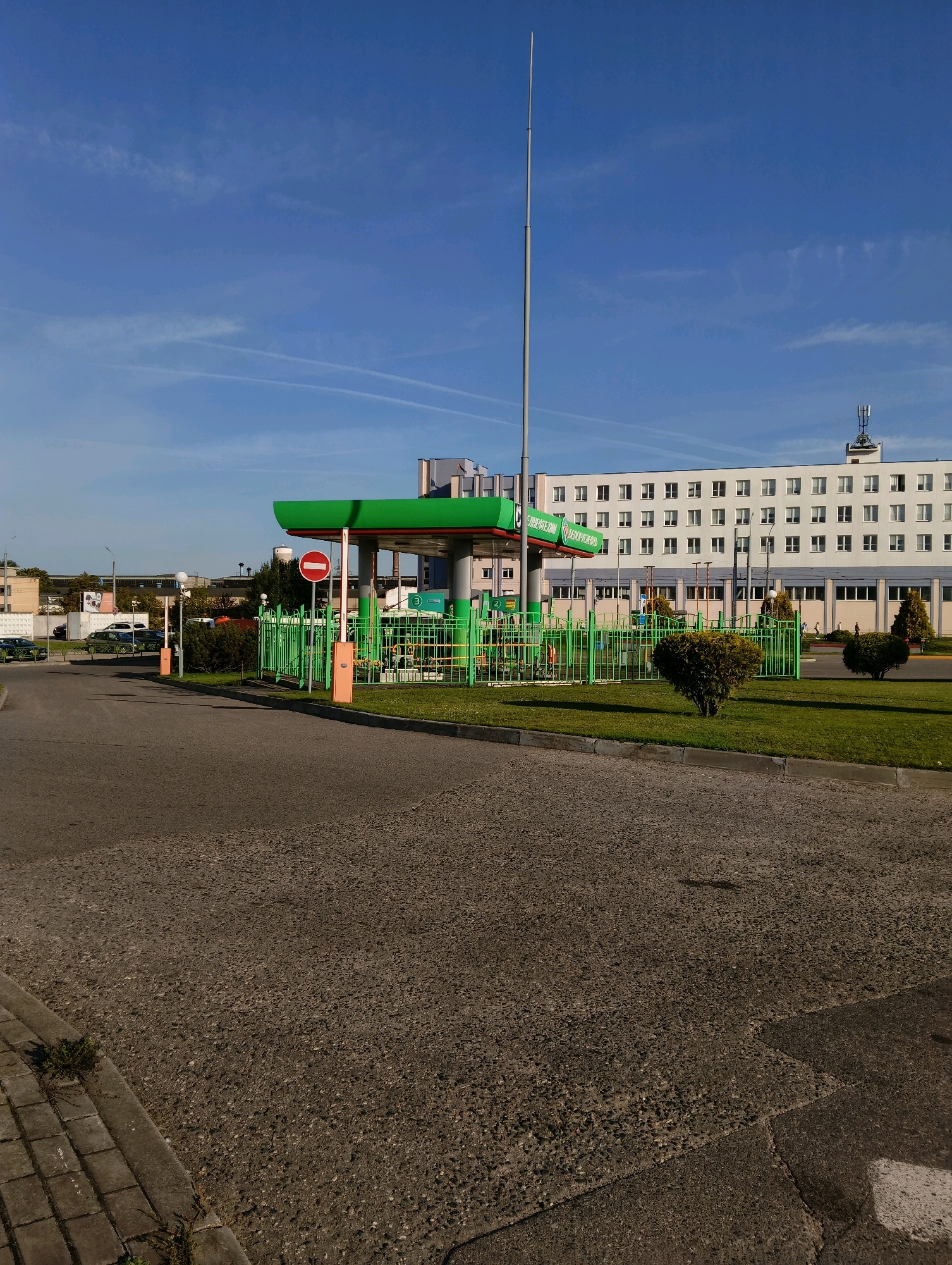 This image depicts a gas station in Hrodna, Belarus. The gas station features a prominent green canopy with red trim, supported by white columns. Several fuel pumps are visible under the canopy, along with a green metal fence surrounding the station. A red and white "no entry" sign is posted on an orange pole. In the background, a long, white building with numerous windows stretches across the scene. A tall, slender radio tower stands against a clear blue sky with faint contrails. The ground in the foreground is paved with asphalt and cobblestones. The overall scene suggests a typical urban environment on a clear day.