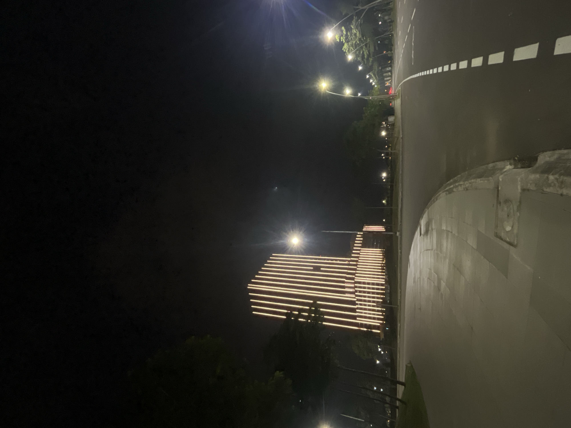 The image depicts an outdoor scene at night in Kosambi Barat, Indonesia. The primary focus is on a modern building illuminated with horizontal bands of warm-toned lights, creating a striking visual against the dark sky. The building appears to be multi-story with a stepped or terraced design, enhanced by the linear lighting.
To the right of the building, a paved road is visible, distinguished by white dashed lines indicating lanes. A sidewalk or pedestrian path runs alongside the road, separated by a raised curb. Streetlights with bright, glowing bulbs are spaced along the side of the road, casting light and contributing to the nighttime ambiance. Trees with dark foliage are scattered throughout the scene, some obscuring parts of the building and road.
The overall impression is one of urban nighttime activity, possibly a commercial or public space given the architecture and road infrastructure. The lighting suggests the scene is well-maintained and actively used, even after dark. The time of day is clearly night, indicated by the darkness of the sky and the artificial illumination. The weather appears clear, with no signs of rain or fog. There is no visible text in the image. The image is taken from a slightly elevated perspective, looking down on the road and towards the building.