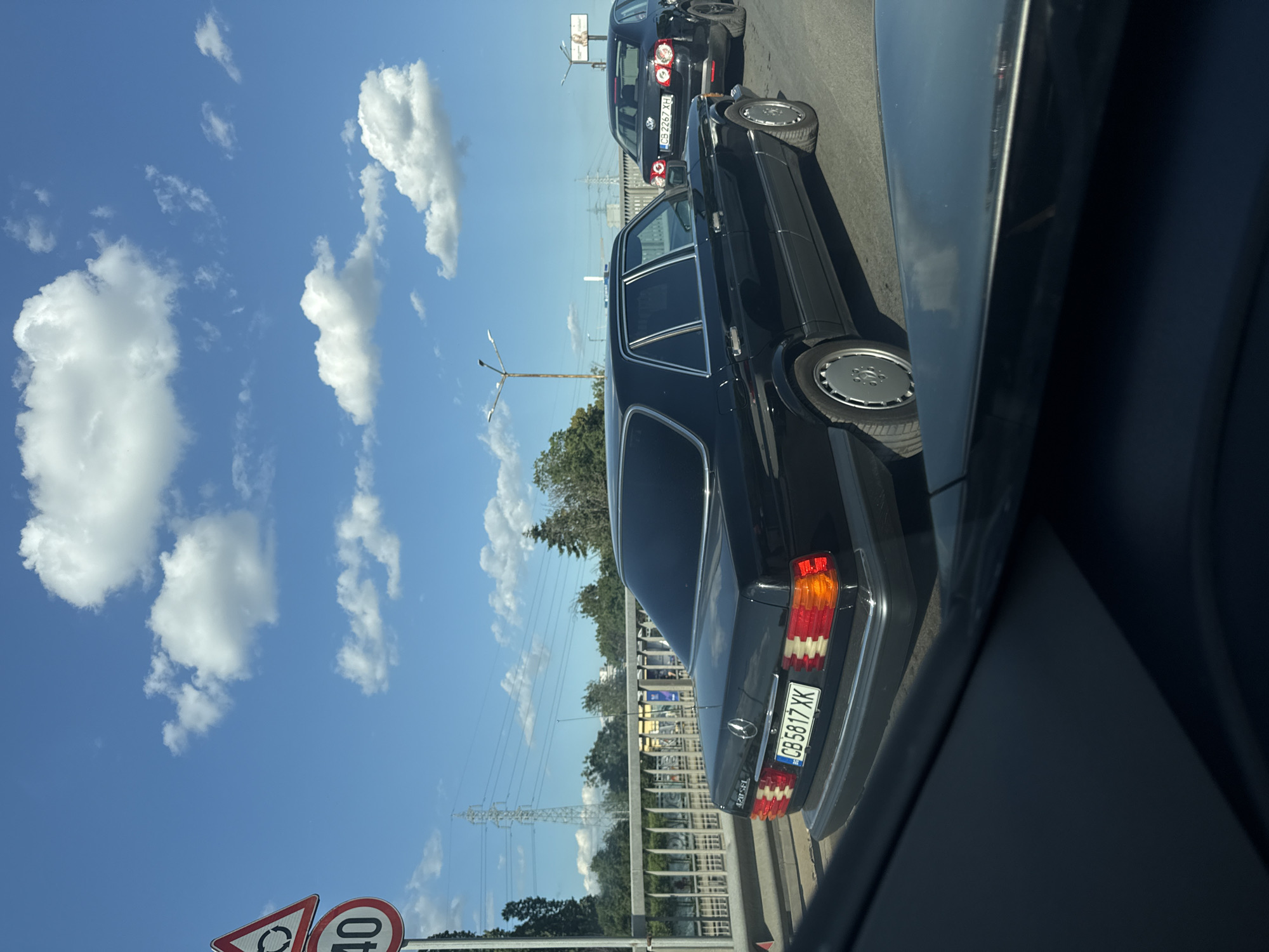 In Sofia, Bulgaria, a black Mercedes-Benz W126 sedan is captured in traffic on a sunny day. The car, bearing the license plate CB 5817 XK, is positioned on a bridge or overpass. Behind it, a black Volkswagen Passat with license plate CB 2267 XH is visible. The sky is blue with scattered white clouds. Road signs are present, including a circular intersection sign and a speed limit sign indicating 40. Electrical pylons and trees line the background. The perspective suggests the photo is taken from another vehicle.