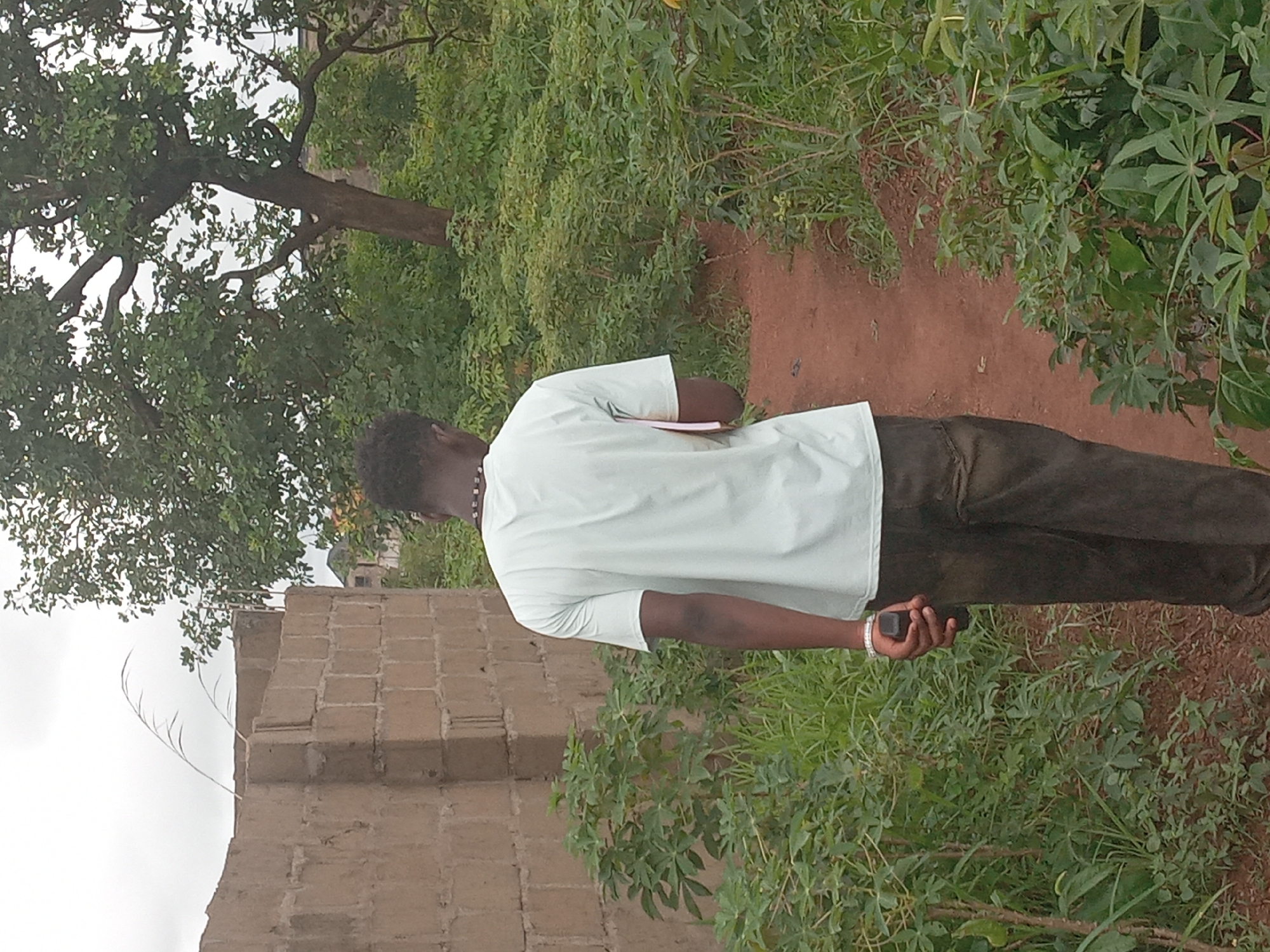 A man stands on a dirt path surrounded by green vegetation in Achara Ndiagu Omegio, Nigeria. He is wearing a light-colored shirt and dark pants, and is holding a small dark object in his right hand. A partially constructed brick wall is visible on the left side of the frame, and a tree with dense foliage is present in the background. The overall scene appears to be outdoors and somewhat rural.