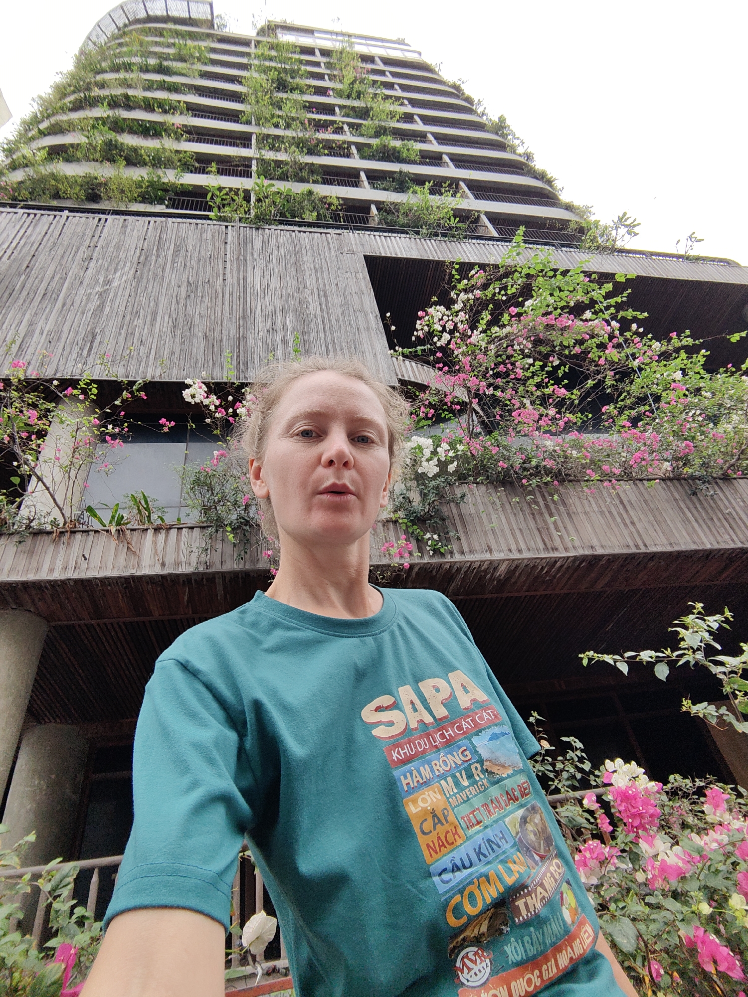 A person is standing in front of a building in Bai Chay Ward, Vietnam. The building features wooden cladding and balconies with plants and flowers. The person wears a green t-shirt with text related to Sapa tourist destinations. The building appears to be a hotel or apartment complex with a rounded, tiered design.