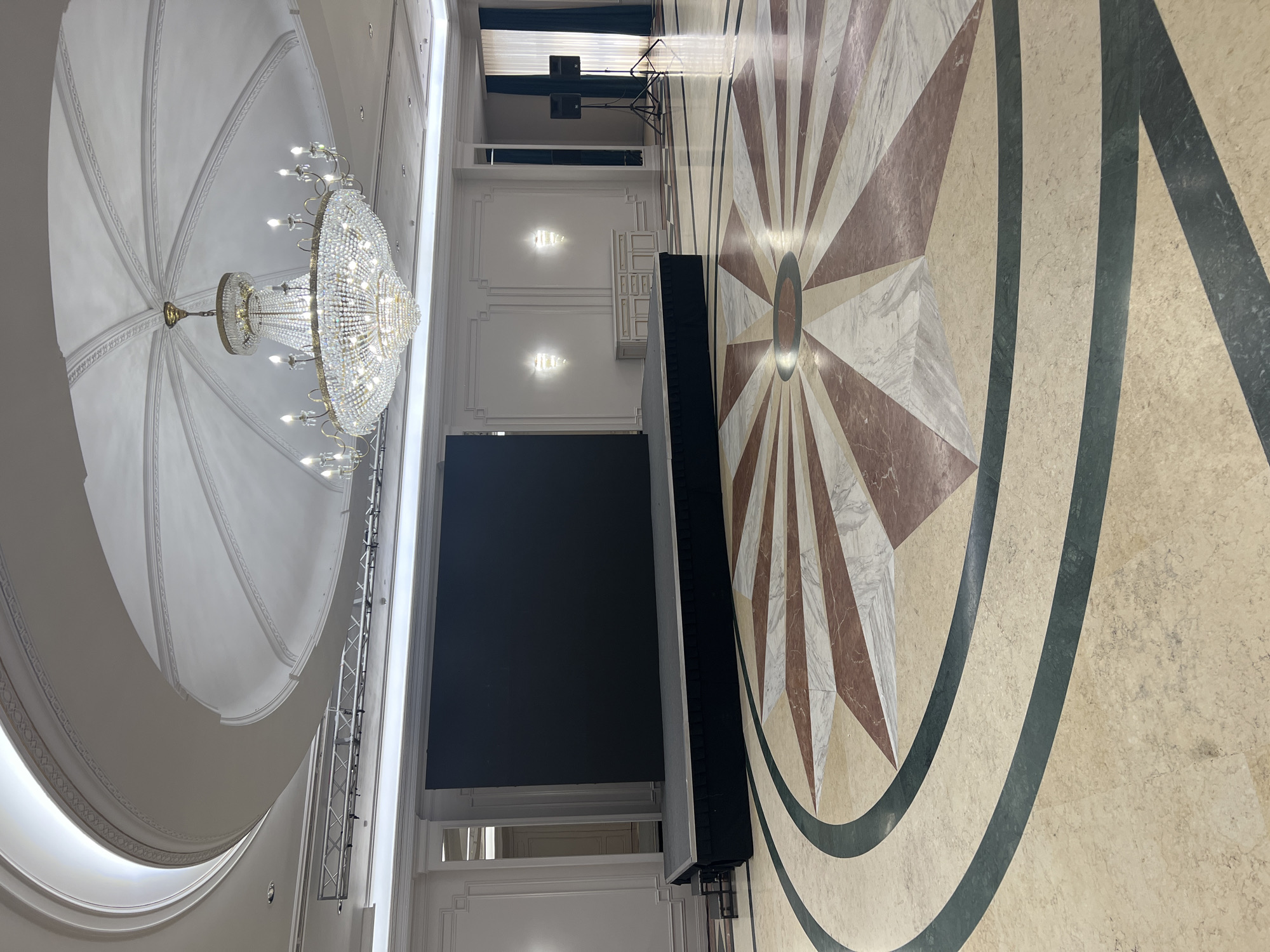 The image depicts an empty event hall, likely in Astana, Kazakhstan, prepared for a gathering. In the foreground, a large, ornate floor mosaic features a sunburst pattern with alternating shades of cream, burgundy, and white marble. Dark green marble bands form concentric circles around the central design.
Dominating the center of the hall is a raised stage covered with dark fabric skirting. Atop the stage rests a massive, dark, rectangular LED screen, currently displaying a solid black image. To the right of the stage, two black speakers on tripods are positioned near a window with dark blue curtains.
The walls are a neutral color, adorned with decorative molding and two illuminated wall sconces. A white credenza with drawers sits against the wall beneath the sconces. The ceiling is a grand, domed structure with intricate white molding and recessed lighting. A large, crystal chandelier hangs prominently from the apex of the dome, its many lights aglow. A metal truss system is visible suspended from the ceiling above the stage, likely for lighting or sound equipment. The overall impression is of a sophisticated and well-appointed venue awaiting an event.