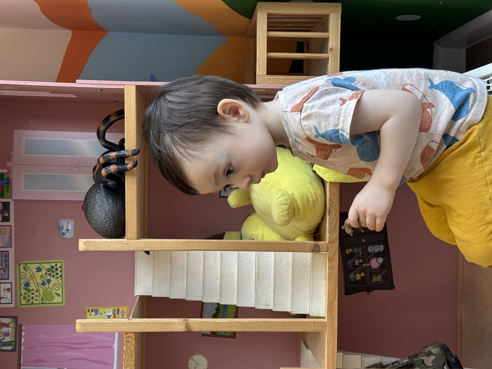 A toddler is reaching into a wooden dollhouse. The dollhouse contains a yellow stuffed animal, a black and orange spider toy, and other miniature objects. The child is wearing a t-shirt with whale and pitcher designs and yellow pants. The background includes a multi-colored wall and a small wooden shelf. The image does not provide any information about the location in the city of Izhevsk, Russia.