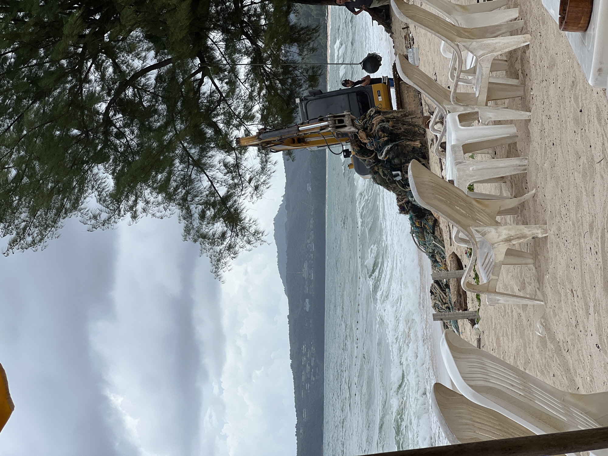 This image depicts a scene on a beach in Choeng Thale, Thailand, characterized by overcast skies and choppy ocean waves. In the foreground, several white plastic chairs and a table are arranged on the sand, suggesting a temporarily abandoned outdoor seating area. To the right of the chairs, a yellow excavator is positioned on the beach, entangled with a large mass of netting and ropes. A dark spherical object hangs from a rope suspended from a tree above the excavator. Two individuals are visible near the excavator; one man in a black t-shirt and cap stands with his arms crossed, facing the excavator, while another man in a grey t-shirt stands further to the right, looking towards the ocean. In the background, the ocean stretches towards a hilly, tree-covered coastline with buildings dotting the slopes. The overall atmosphere is one of disruption and ongoing work, possibly related to clearing debris from the beach or ocean.