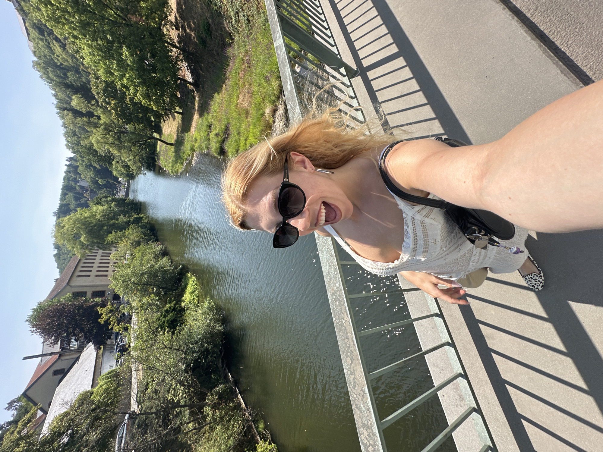 A young woman with blonde hair and sunglasses is taking a selfie on a bridge overlooking a river in Vianden, Luxembourg. She is wearing a white lace dress and animal print shoes. The river is surrounded by lush green trees and buildings. The sun is shining, casting shadows on the bridge.