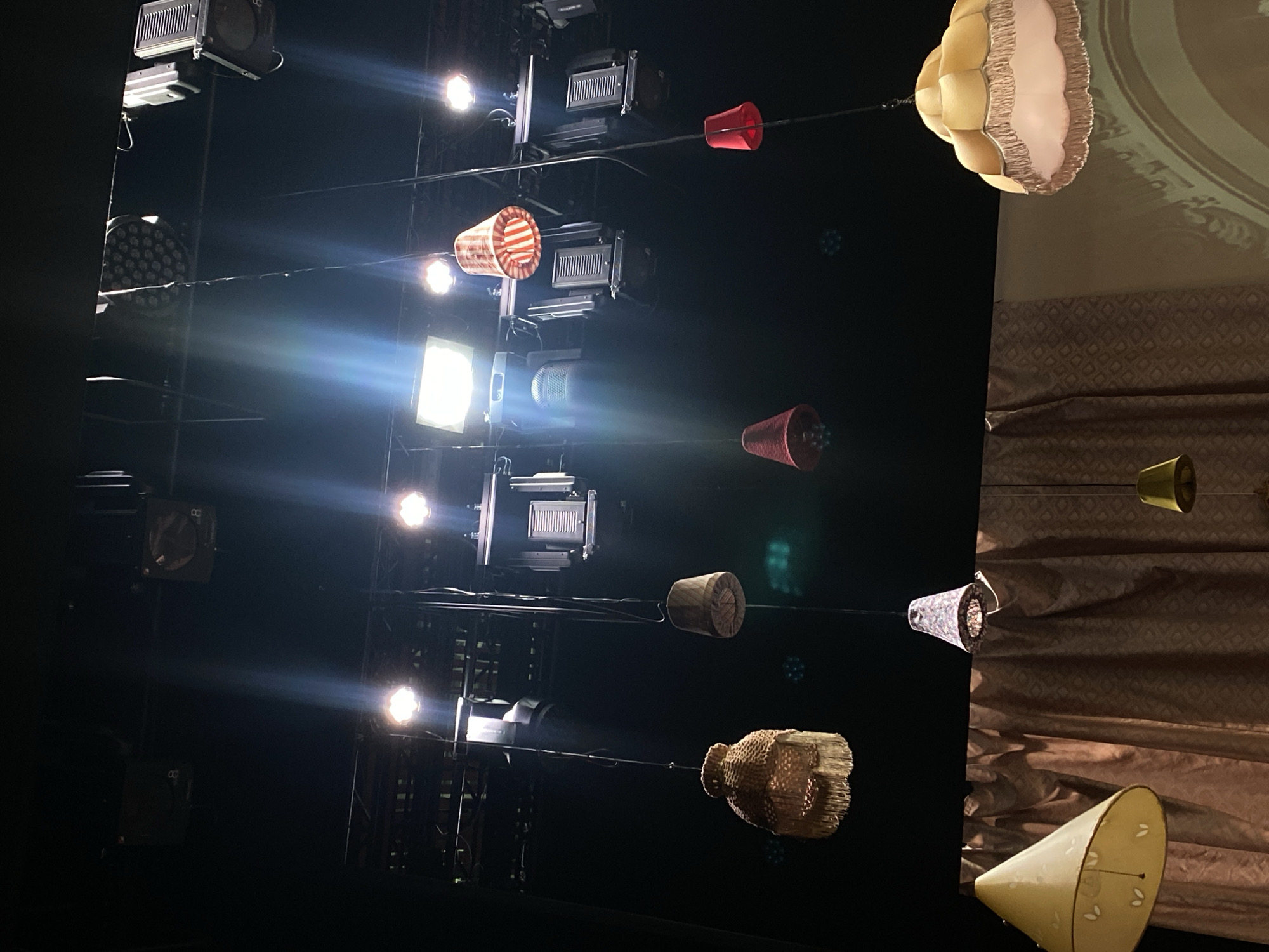 The image shows a view upward towards a stage lighting rig and decorative lighting fixtures, possibly in a theatre or event space in Zelenograd, Russia. The lighting rig on the left comprises several black metal structures holding various spotlights and lighting panels, some of which are emitting bright beams of light. Wires are strung across the frame, suspending a series of decorative lampshades in various shapes, sizes, and colors, including red, striped red and white, wood-like, woven gold, blue with a floral pattern, and cream. The background to the right shows patterned wallpaper and a section of draped fabric in brown tones with a subtle geometric pattern.