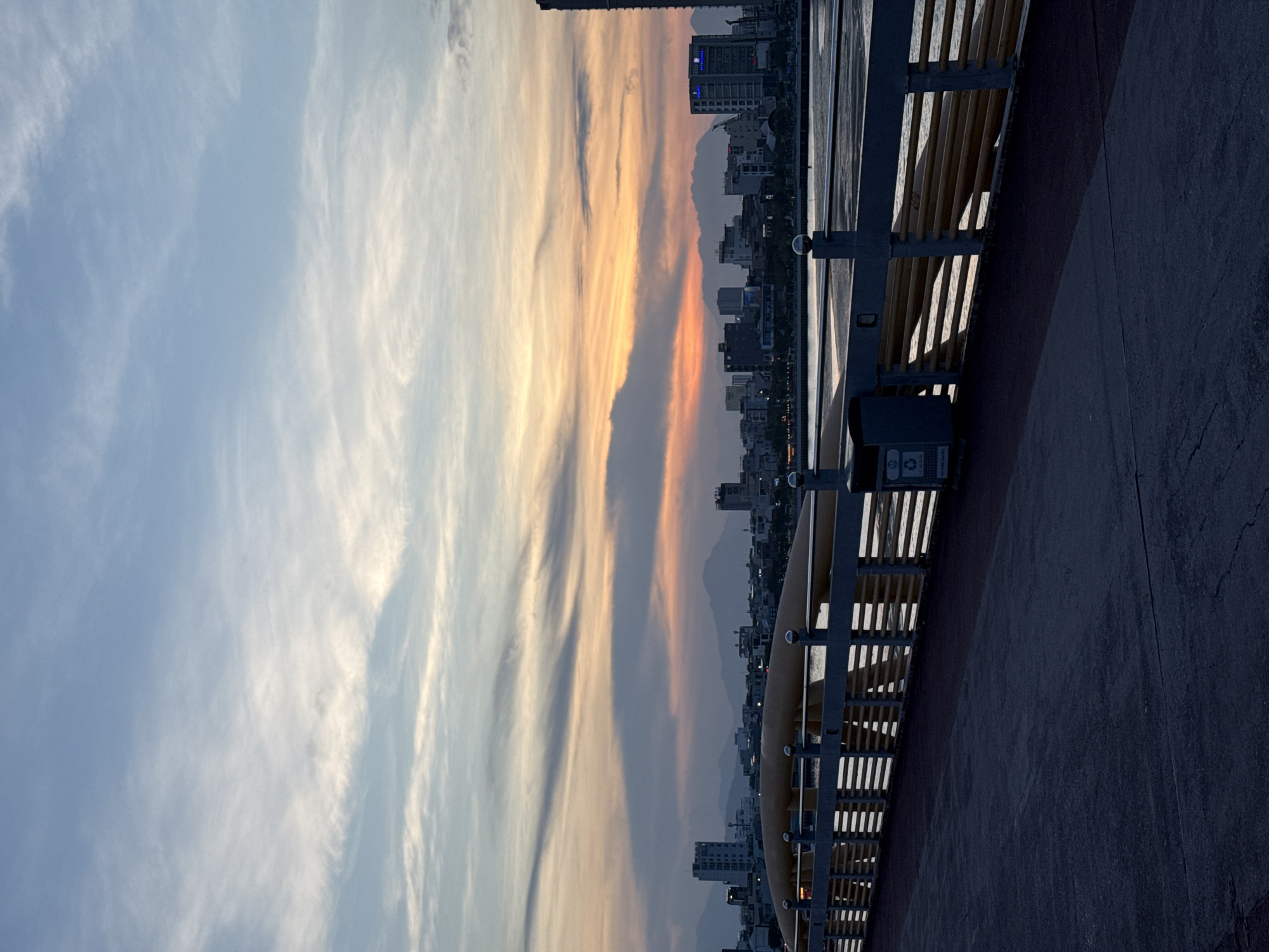 The image depicts a view from a bridge overlooking the city of Da Nang, Vietnam, at dusk. The sky is a canvas of soft blues and oranges, with wispy clouds streaking across the horizon. In the distance, a cityscape of buildings, including high-rises, stretches along the coastline, with mountains forming a backdrop. The bridge itself has a distinctive curved railing and a dark paved walkway. A dark trash receptacle with recycling symbols is visible on the right side of the walkway. The water below the bridge appears calm, reflecting the muted colors of the sky. There are no people or animals visible in the frame. The overall atmosphere is serene and picturesque, capturing the transition from day to night in the coastal city.