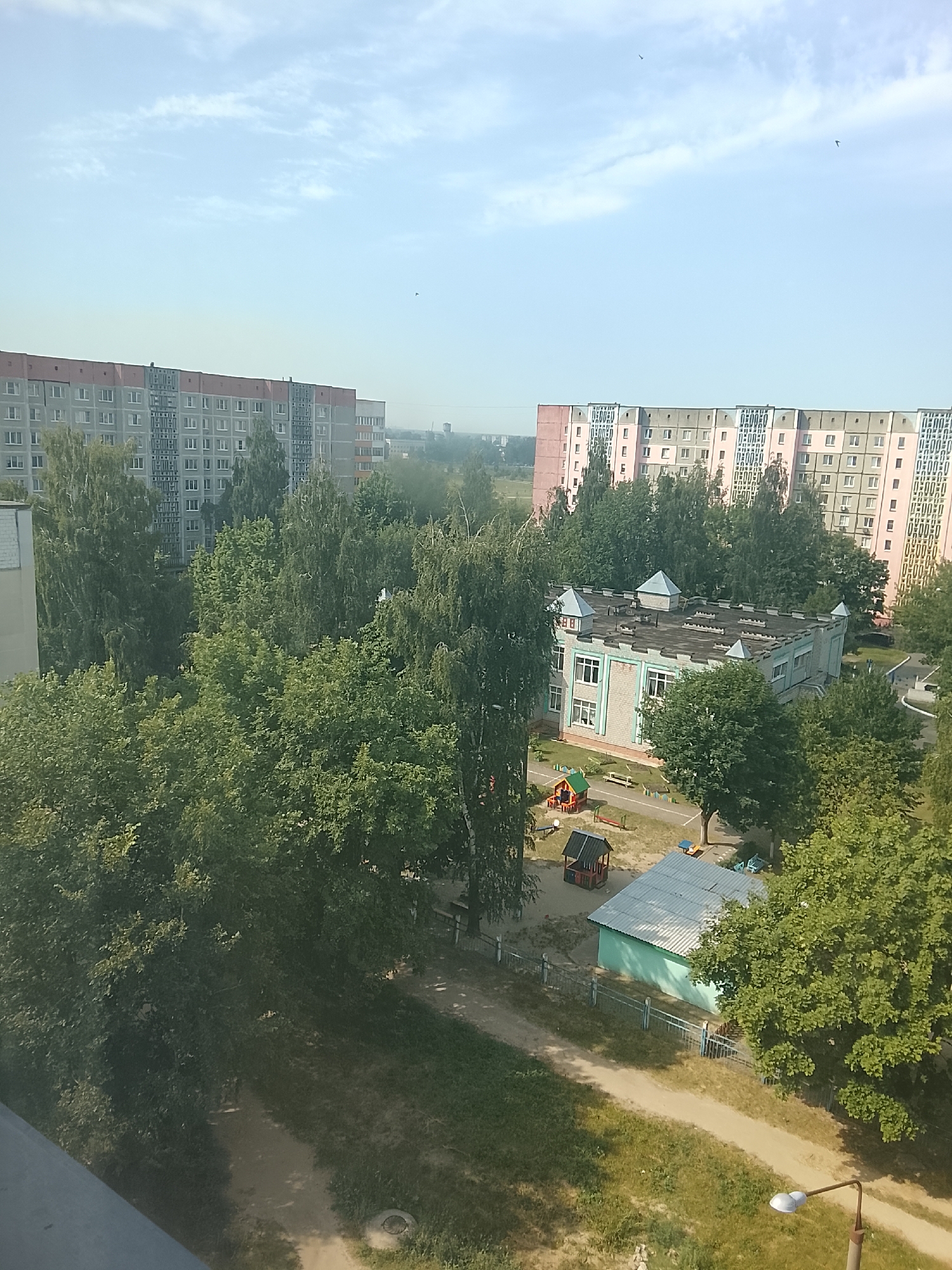 The image is an aerial view of a residential area in Zhlobin, Belarus, on a sunny day. In the foreground, a dirt path winds through grassy areas with mature trees. A modern street lamp with two lights is visible on the right. In the middle ground, a small, light blue building with a metal roof, possibly a shed or utility structure, is fenced off. Adjacent to this is a playground with colorful climbing structures and a small wooden house. Further back, a two-story building with a castle-like facade and turquoise trim, likely a kindergarten or pre-school, stands among the trees. In the background, two large apartment buildings dominate the skyline. The left building is gray with a red-orange band under the roof. The right building is pink and green with repeating window patterns. The sky is a pale blue with scattered wispy clouds and two birds flying. The overall scene depicts a typical urban environment with residential buildings, green spaces, and recreational areas.