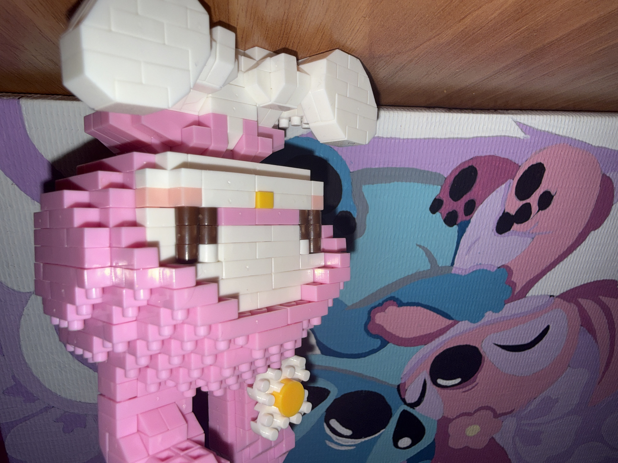 The image shows a close-up of a pink bunny-shaped toy made of interlocking plastic bricks. The toy is sitting on a wooden surface. In the background, there is a canvas print featuring cartoon characters resembling Stitch and Angel from the Lilo & Stitch franchise. The characters are depicted in pink and blue colors. The overall scene appears to be indoors. There is no information available to indicate the location as Rayevskiy, Russia.