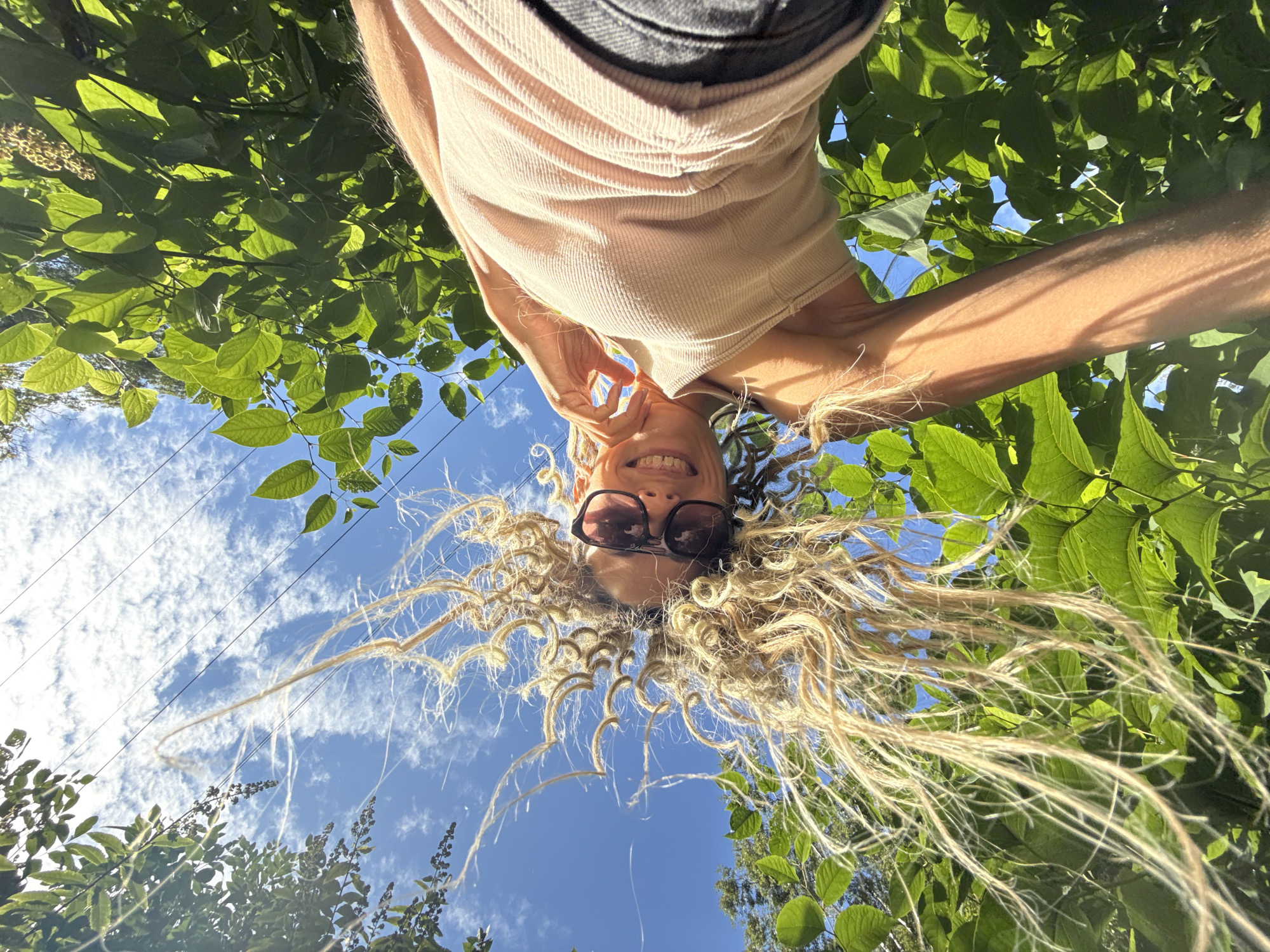 In Fedorino, Russia, a woman with blonde dreadlocks and sunglasses is captured in a low-angle shot, looking up at the camera. She is surrounded by lush green foliage, with a bright blue sky dotted with clouds visible above. She wears a light-colored ribbed tank top and dark pants. Her hair is spread out, catching the light and creating a dynamic visual effect. The sun illuminates her face and arm, casting shadows through the leaves. Power lines are visible in the upper portion of the frame, cutting across the sky.
