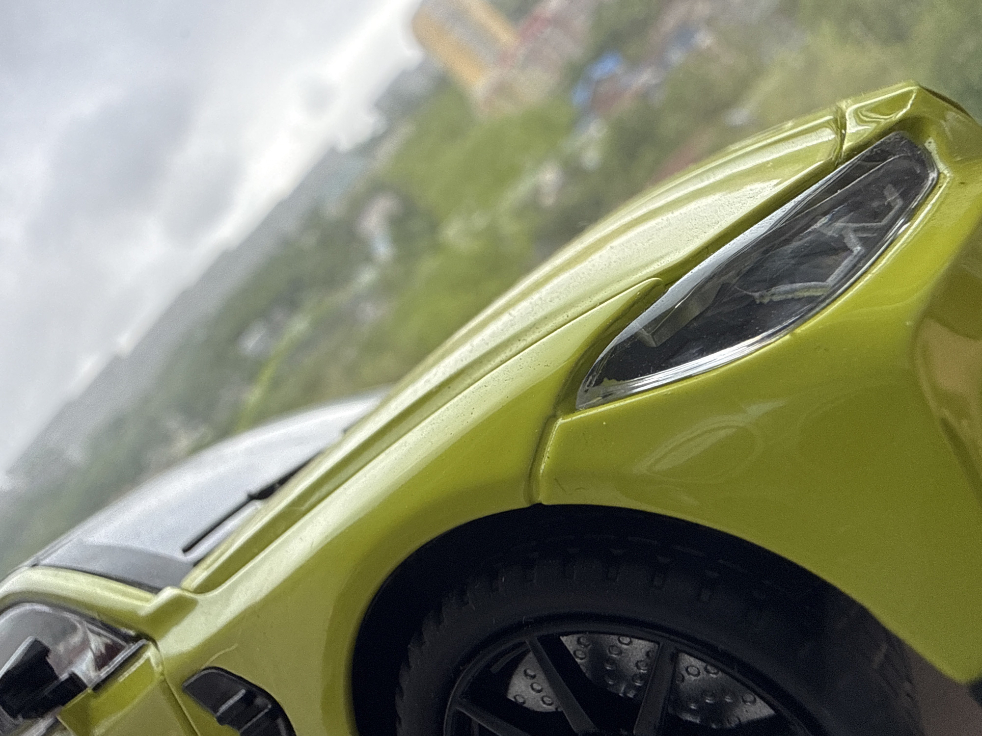 A close-up shot of a lime-green toy car is positioned in front of a blurry background. The background shows a city scene with buildings, greenery, and overcast skies. It is impossible to verify the location is Vladivostok. The toy car is the main subject. The focus is on the car's headlight, front fender, and wheel. The wheel is black with visible brake discs. The car has a silver hood. The overall composition is tilted. The car shows minor imperfections and what appears to be water droplets on its surface.
