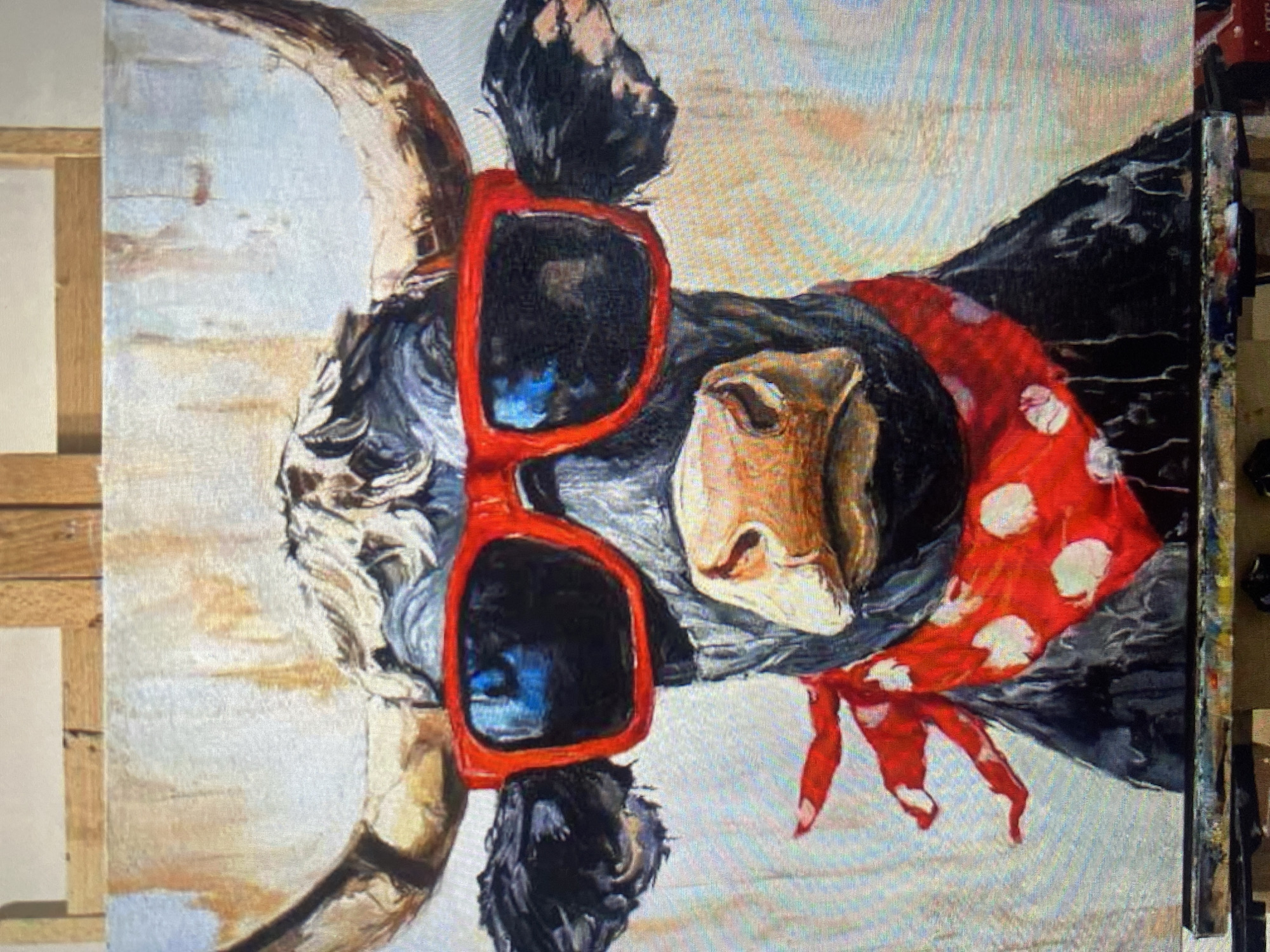 A painting on canvas depicts a black bull wearing red sunglasses and a red and white polka dot bandana. The bull has large horns and is positioned in the center of the frame. The background is abstract with light and dark brushstrokes. The painting is on an easel, with wooden beams visible above it. The bottom of the frame shows a palette with various colors of paint and brushes. The location is unknown, in the city of Unknown, Unknown.
