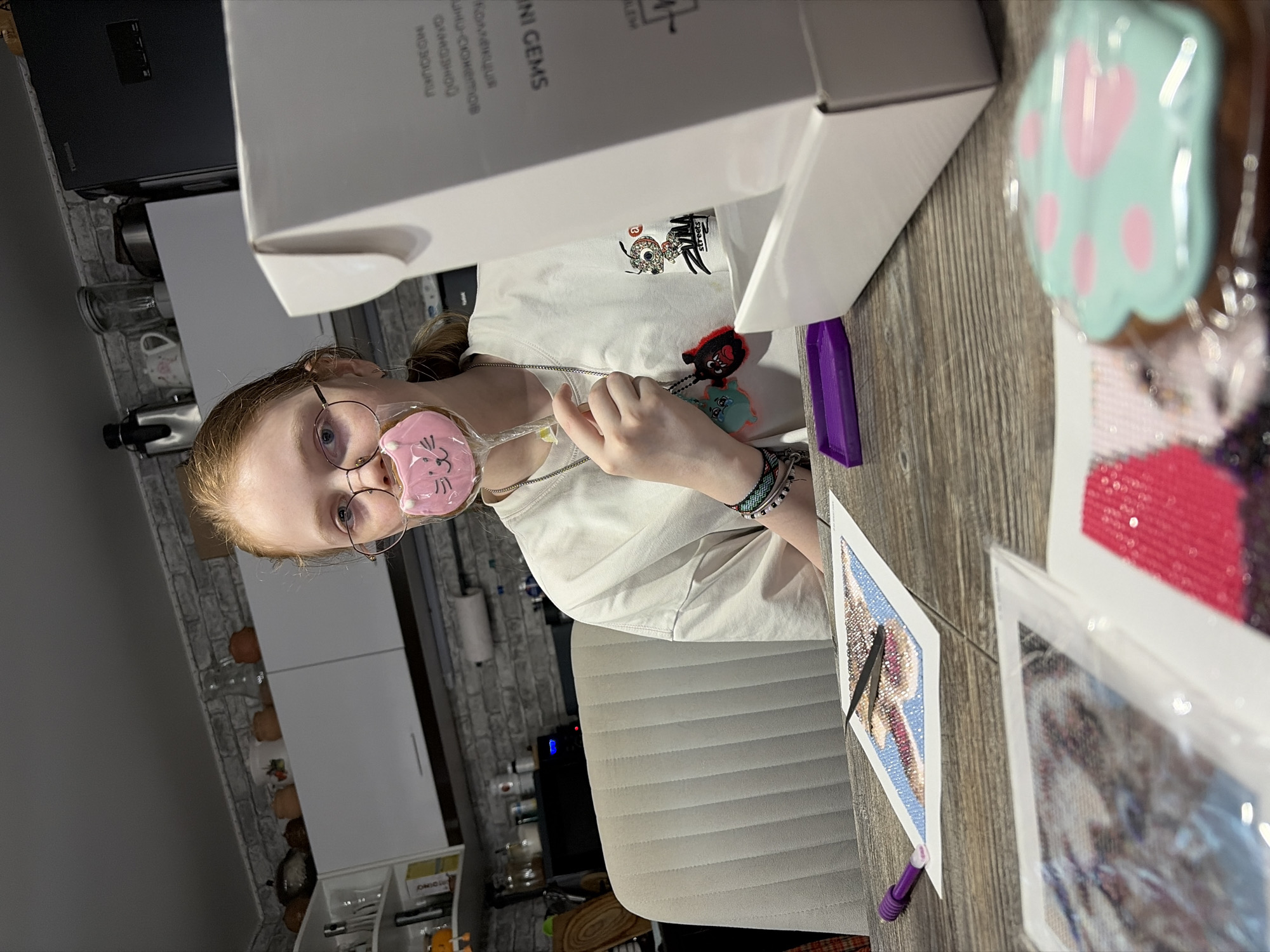 A young woman sits at a wooden table in what appears to be a home in Chaikovsky, Russia. She is facing the camera and holding a pink lollipop shaped like a cat's face in front of her left eye. The woman wears round-framed glasses, a white t-shirt with an image on it, and several bracelets on her left wrist. In front of her on the table are diamond painting projects and a purple tray. To the left of the frame is a partial view of a kitchen, with white cabinets and a gray brick wall. A light gray chair is visible in the background. The box on the table is labeled "MINI GEMS" in Russian.