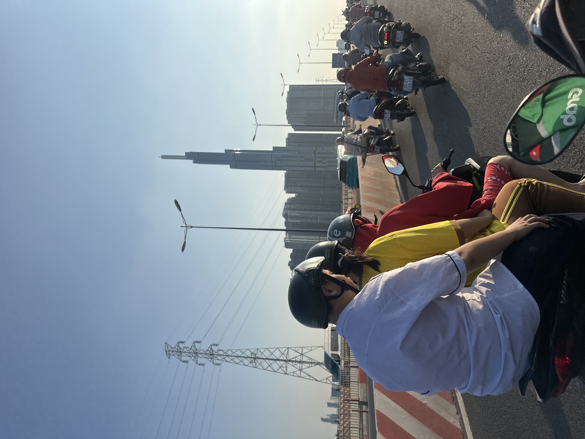The image shows a group of people on motorbikes on a bridge in Thủ Đức, Vietnam. The riders are wearing helmets and casual clothing, with at least 3 of them sitting closely together. Some motorbikes are moving slowly in traffic. In the background, the Landmark 81 skyscraper is visible among other buildings and overhead power lines. A "Grab" logo is visible on a mirror. The sky is clear and bright.