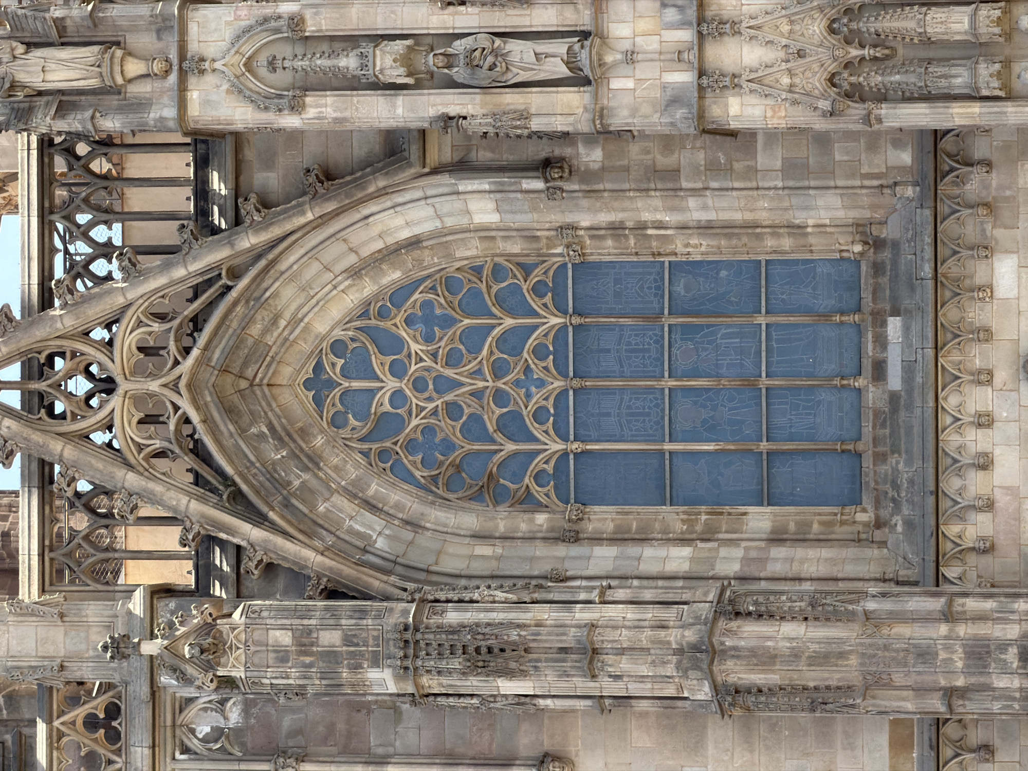 This is a close-up, eye-level, vertical photograph of a Gothic cathedral facade in Barcelona, Spain. The building is constructed of weathered, beige stone blocks. The focal point is a large, arched stained-glass window with a complex tracery pattern at the top. The lower portion of the window is divided into rectangular panes with faint, etched depictions of figures and architectural elements, likely religious in nature. The blue of the stained glass is muted, suggesting it is old or the light is indirect.
Flanking the window are prominent stone architectural features. To the left, a tall, fluted column with intricate Gothic carvings and gargoyles is visible. To the right, a niche contains a standing statue of a robed female figure holding a book or small object. Above and around the main window, more elaborate stone carvings, pinnacles, and decorative arches are present, indicative of Gothic architecture.
Below the window, a decorative band of small, pointed arches supported by columns runs across the facade, with small carved faces visible within some of the arches. The stone appears aged and somewhat worn. The sky is a pale blue, visible at the very top of the image through openings in the upper architectural details. The image captures a section of the facade of the Barcelona Cathedral, specifically the west front.