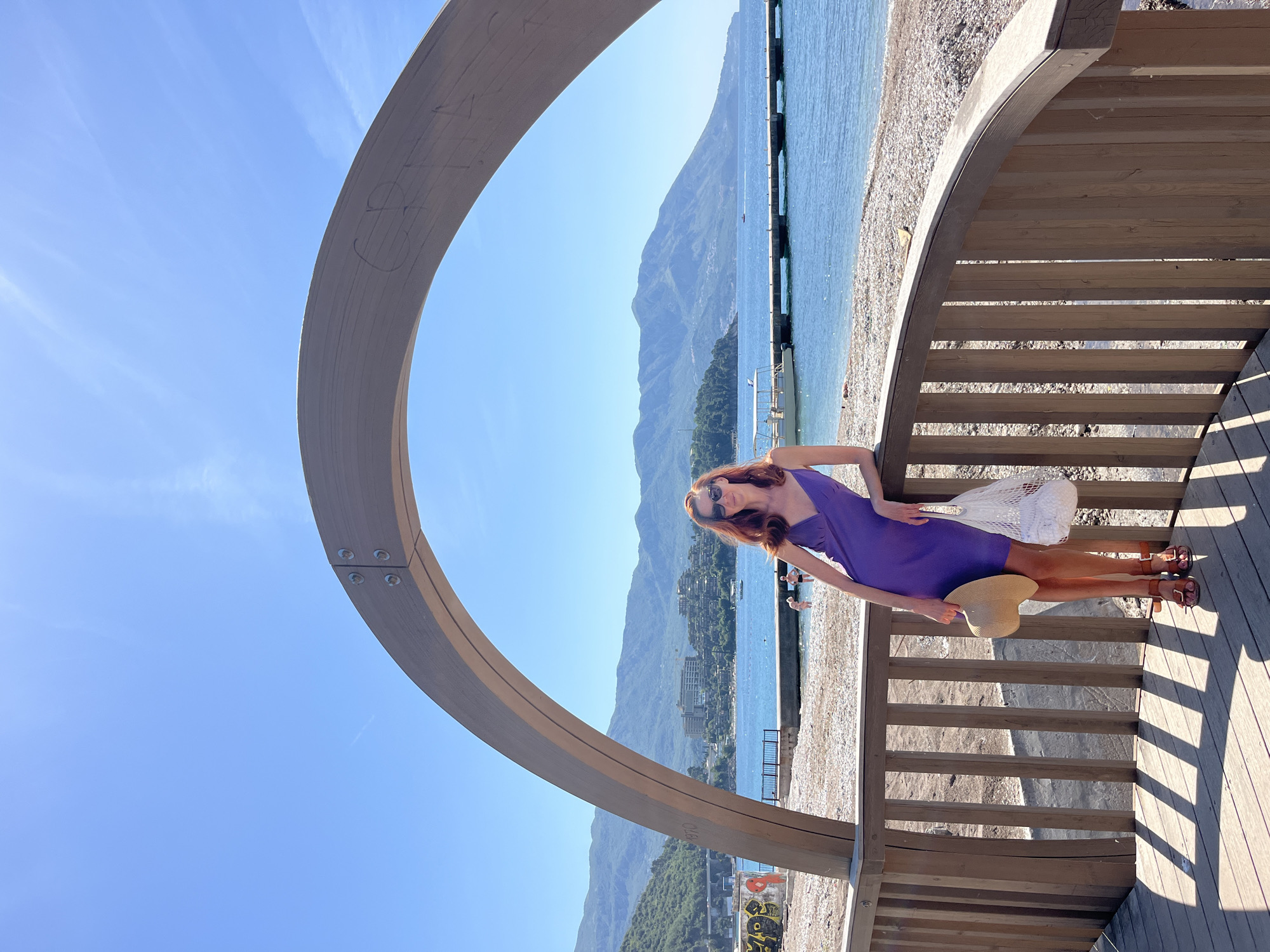 A woman with reddish-brown hair stands on a wooden platform, leaning against a wooden railing and structure, wearing a purple dress and holding a straw hat in her left hand, with a white mesh bag at her side. The woman is facing the right of the frame, partially framed by a wooden arch. She is wearing sunglasses. Beyond the structure and railing is a beach with a pebble surface and calm sea water. In the background, mountains are visible on the opposite side of the water. The location is Budva, Montenegro.