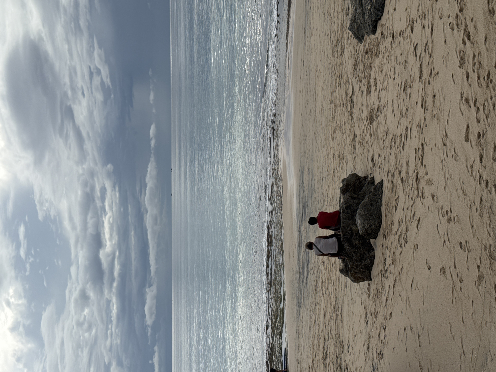 The image captures a serene beach scene in Kuta Selatan, Indonesia, featuring the ocean, sandy shore, and a dramatic sky. In the foreground on the right, two individuals are seated or leaning against dark, jagged rocks on the light beige sand, which is marked with numerous footprints. One person wears a red top, and the other a white top, both appear to be facing towards the expansive ocean.
The sea, occupying the left and central portions of the image, reflects a bright, silvery light, suggesting either early morning or late afternoon sun. Gentle waves break along the waterline, creating a foamy edge where the water meets the sand. The sky above is filled with a thick layer of grey and white clouds, forming interesting and varied patterns, indicating an overcast or partly cloudy day. The horizon line is visible where the ocean meets the sky. The overall atmosphere is calm and contemplative, with the figures seemingly enjoying the peaceful coastal setting. No visible text is present in the image.