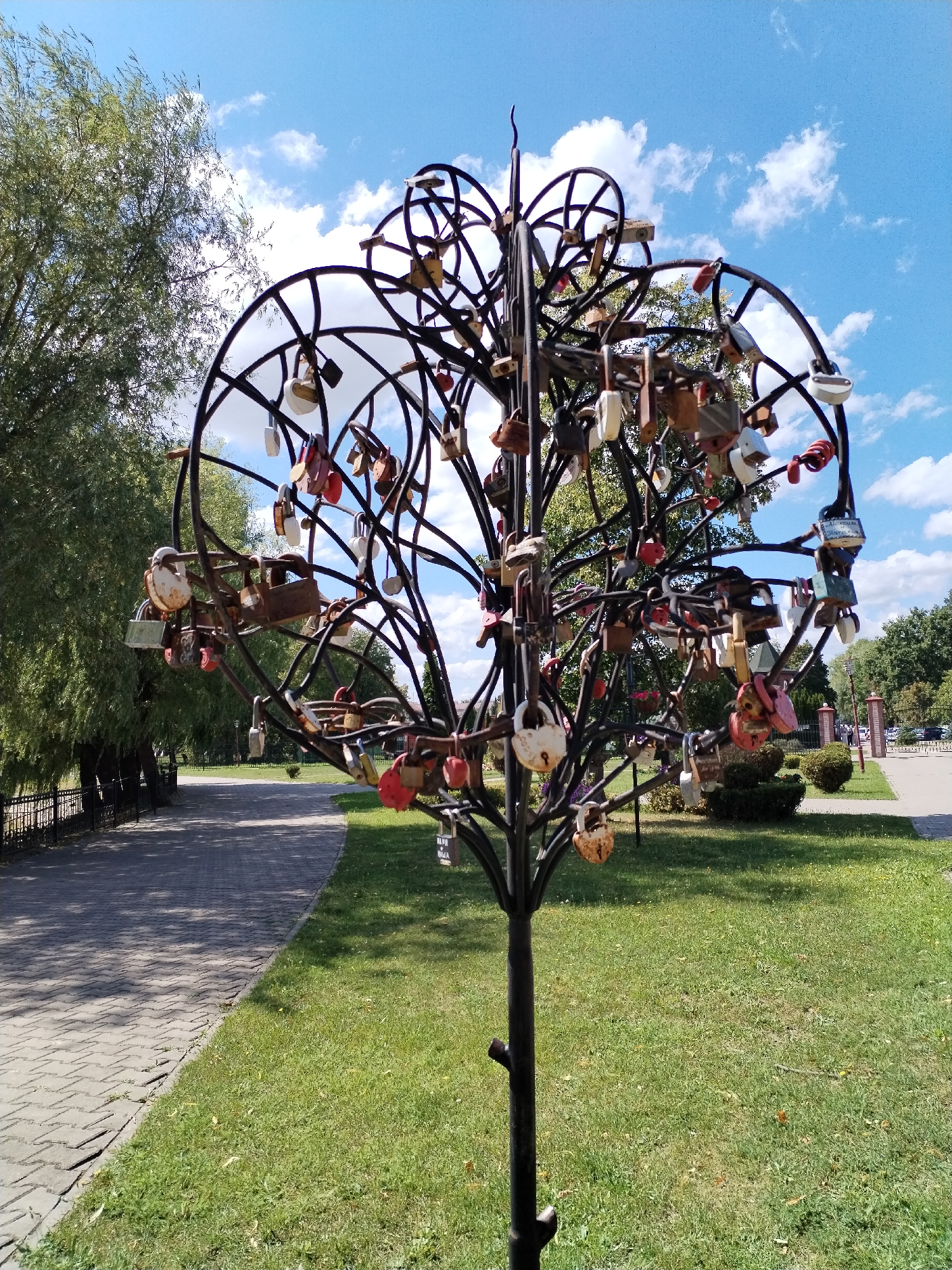 An outdoor, eye-level, medium shot captures a sculpture of a tree adorned with numerous padlocks, set against a backdrop of a park with trees, grass, a paved walkway, and a blue sky with white clouds. The sculpture, made of black metal, features an intricate, branching structure shaped like a heart at the top, with many metal loops extending from it. Attached to these loops are a variety of padlocks in different colors and states of wear, including rust. The tree sculpture stands in a grassy area next to a gray, paved walkway. In the background, more trees line the walkway, and a black metal fence is visible. Beyond the fence, a pathway leads to a pedestrian area with more trees and a building with a fence and columns. The sunlight casts shadows on the grass and walkway, indicating it is daytime. No people are visible in the immediate vicinity of the sculpture.