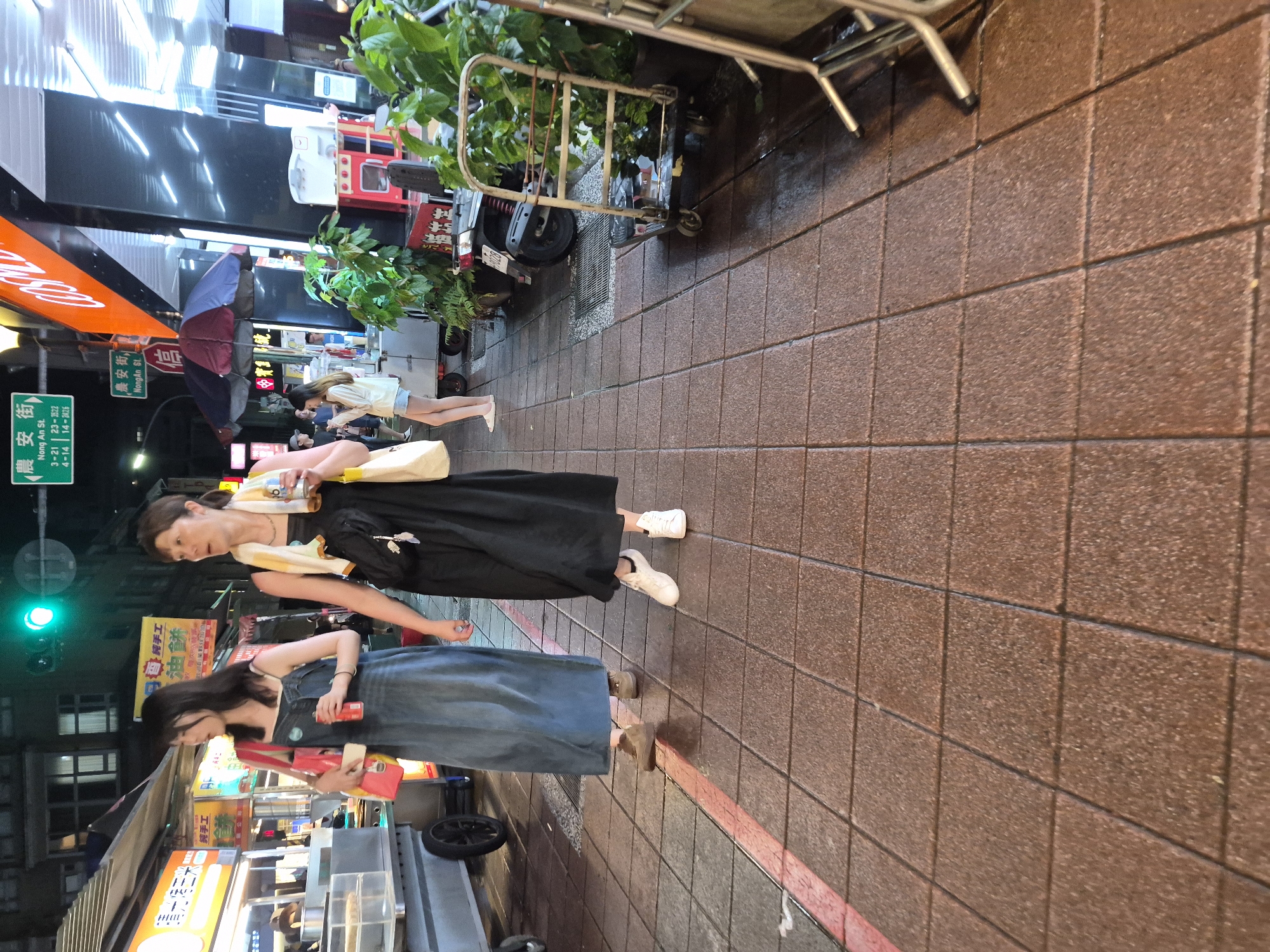 The image captures a street scene at night in Taipei, Taiwan. Two women are walking on a tiled sidewalk. The woman on the left is wearing a denim dress and carrying a pink bag. The woman on the right is wearing a black dress and has a towel draped around her shoulders. In the background, there are street vendors with illuminated signs, a traffic light, and a parked motorcycle. The overall impression is that of a bustling night market or street with ongoing commercial activity.