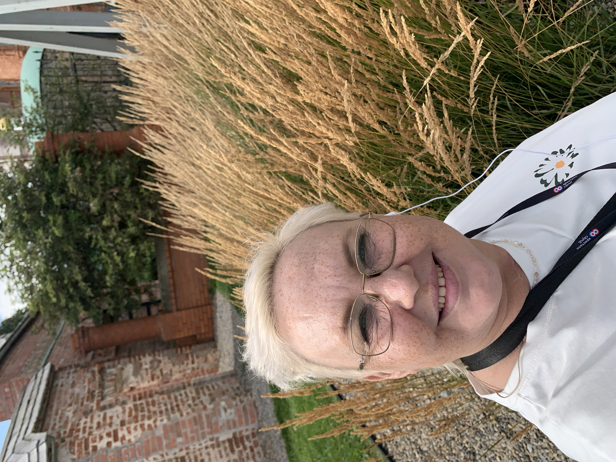 The image shows a close-up, waist-up portrait of a Caucasian woman with short, blonde hair, fair skin with freckles, and glasses. She is wearing a white t-shirt, a black lanyard with a logo that includes a daisy, and white earphones. She is smiling at the camera. Behind her, there is a large patch of dry, golden grass, a brick wall, and some greenery. The scene appears to be outdoors, possibly in a park or garden. The location is specified as Usolye, Russia.