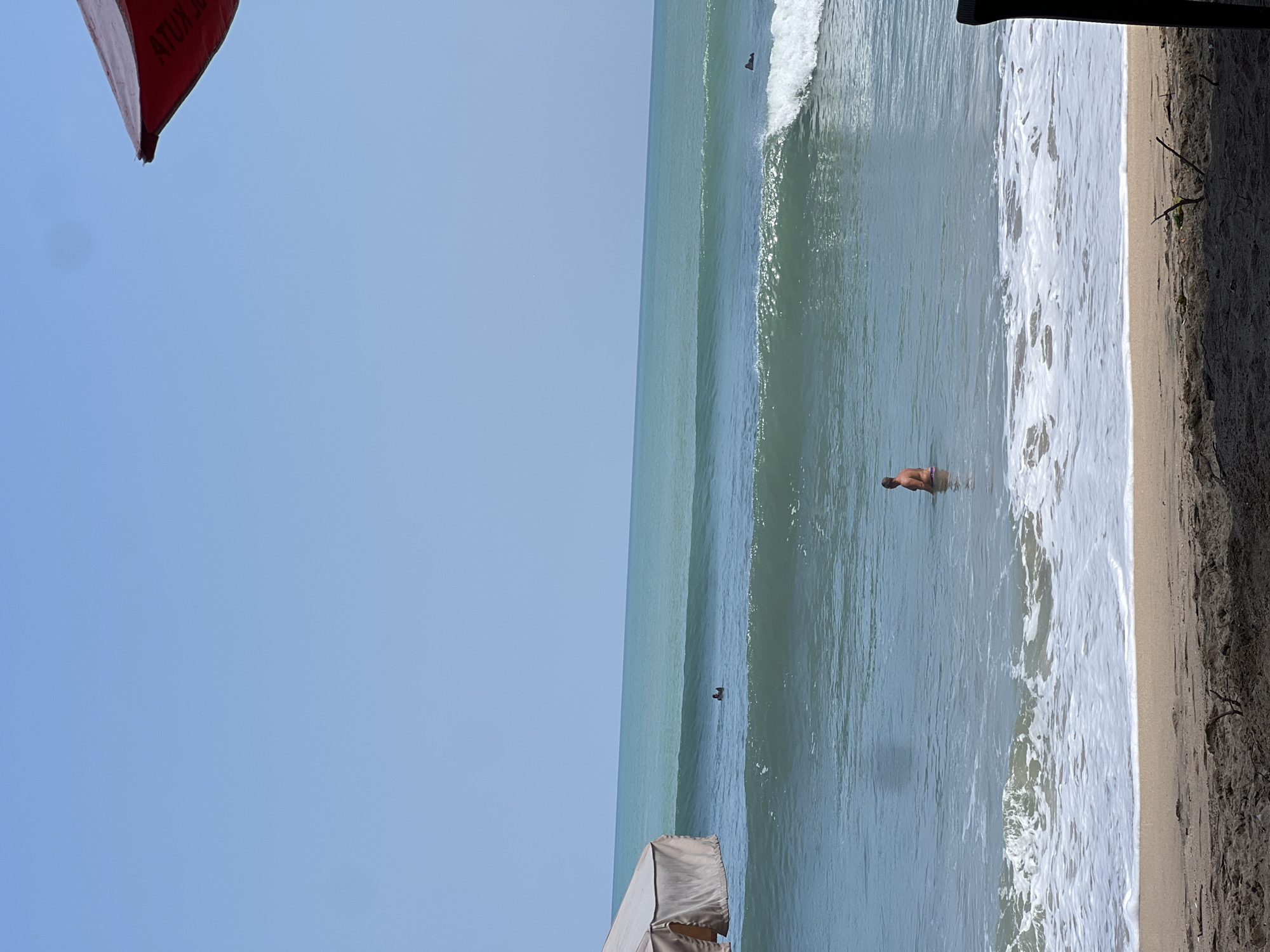 A wide shot captures a beach scene in Kuta, Indonesia, on a clear, sunny day. The ocean, with its characteristic turquoise waves, dominates the frame. In the foreground, the sandy beach transitions to the water, with gentle waves lapping the shore and creating white foam. In the mid-ground, a person stands in the shallow water, facing away from the camera, appearing to be enjoying the ocean. In the background, the vast expanse of the ocean stretches to the horizon, meeting the pale blue sky. On the left side of the image, a part of a tent or canopy is visible, providing shade. The overall atmosphere is one of a tranquil beach day.