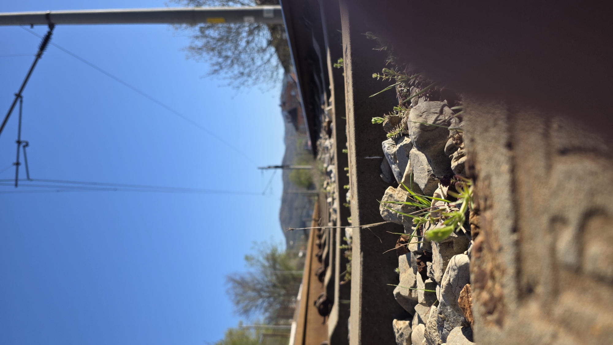 The photograph depicts a close-up view along a railway track, possibly in the Blagoevgrad region of Bulgaria given its mountainous backdrop. The foreground shows the side of a railway track cluttered with small rocks and vegetation. The track itself appears aged, displaying signs of rust and wear. In the background, one sees a blue sky and overhead power lines running parallel to the rails. Further in the distance, there's a glimpse of a mountainous landscape dotted with trees and possibly some buildings. The overall perspective suggests the shot was taken from a low angle, focusing on the textures and details of the immediate surroundings.