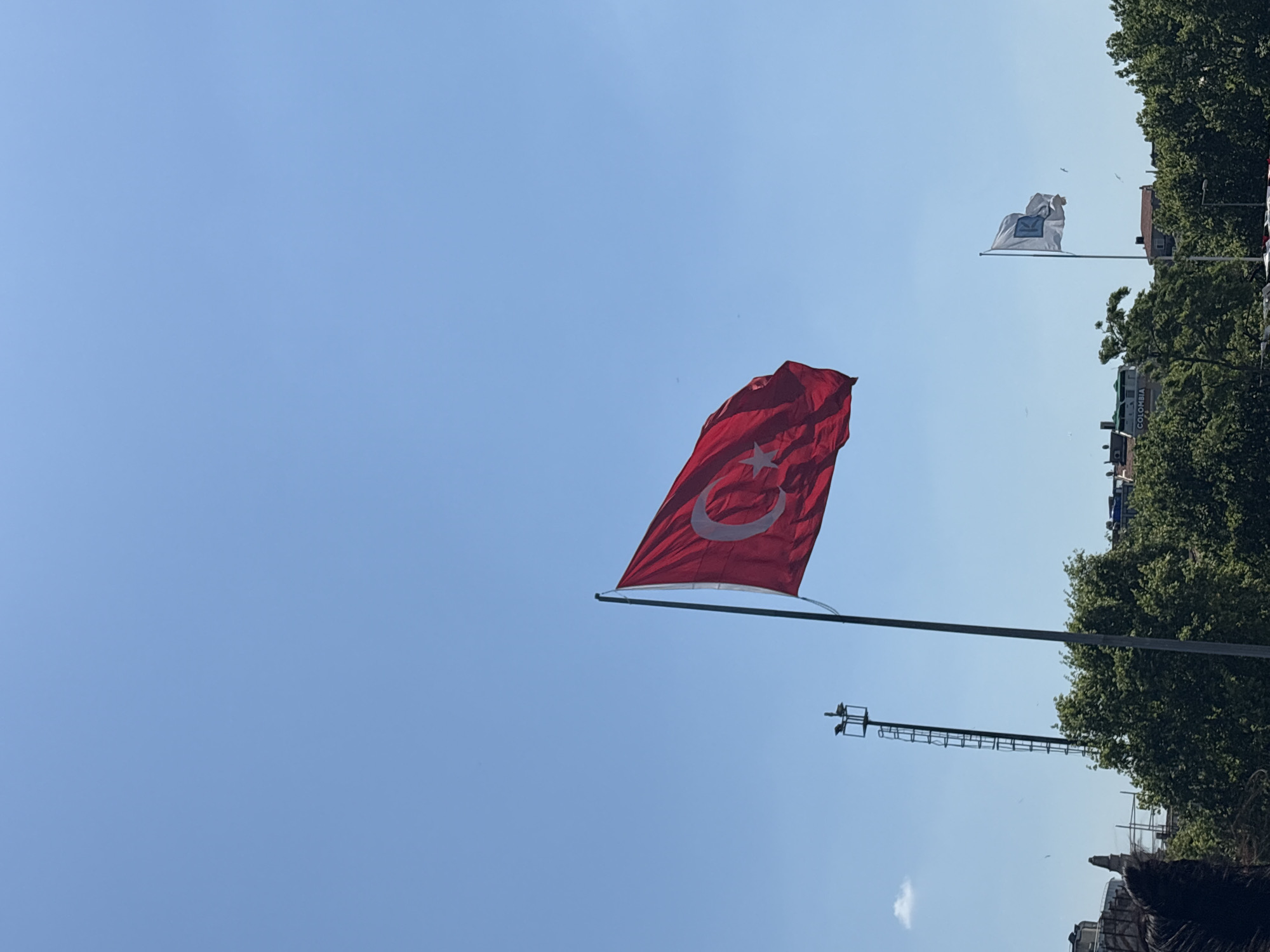 The image depicts a clear blue sky over Kadıköy, Turkey, with a Turkish flag prominently displayed on a flagpole in the foreground. The flag is red with a white crescent and star. To the left and slightly behind the Turkish flag, a tall metal structure with a ladder is visible. In the background, lush green trees form a dense canopy, interspersed with buildings. Another smaller flag, white with a blue emblem that appears to be the letter "K", is flying on a separate flagpole to the right of the Turkish flag. A building with "COLOMBIA" visible on its facade can be seen through the trees. The overall scene suggests an outdoor setting on a sunny day in the city.