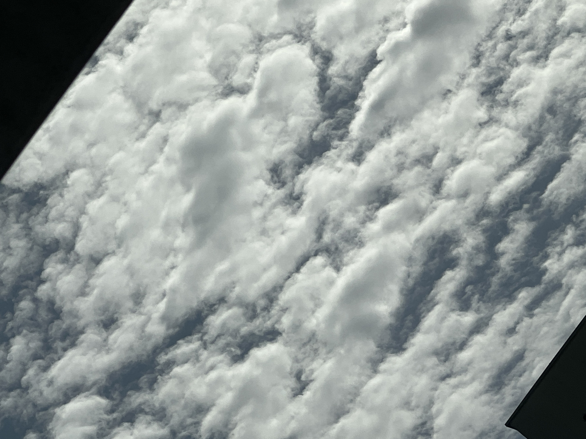 The image shows a close-up view of a cloudy sky, with the clouds appearing to be arranged in parallel rows. The clouds are white and fluffy, with some areas appearing darker and more textured. The sky is a pale blue, and the overall impression is one of a dynamic and somewhat overcast day. There are no people, buildings, or other objects visible in the image, apart from a dark triangular shape in the bottom left corner that could be the edge of a building or another structure, and a dark diagonal line in the top right corner that appears to be a shadow or the edge of an object. The image does not contain any visual elements that would indicate the location in Warsaw, Poland.