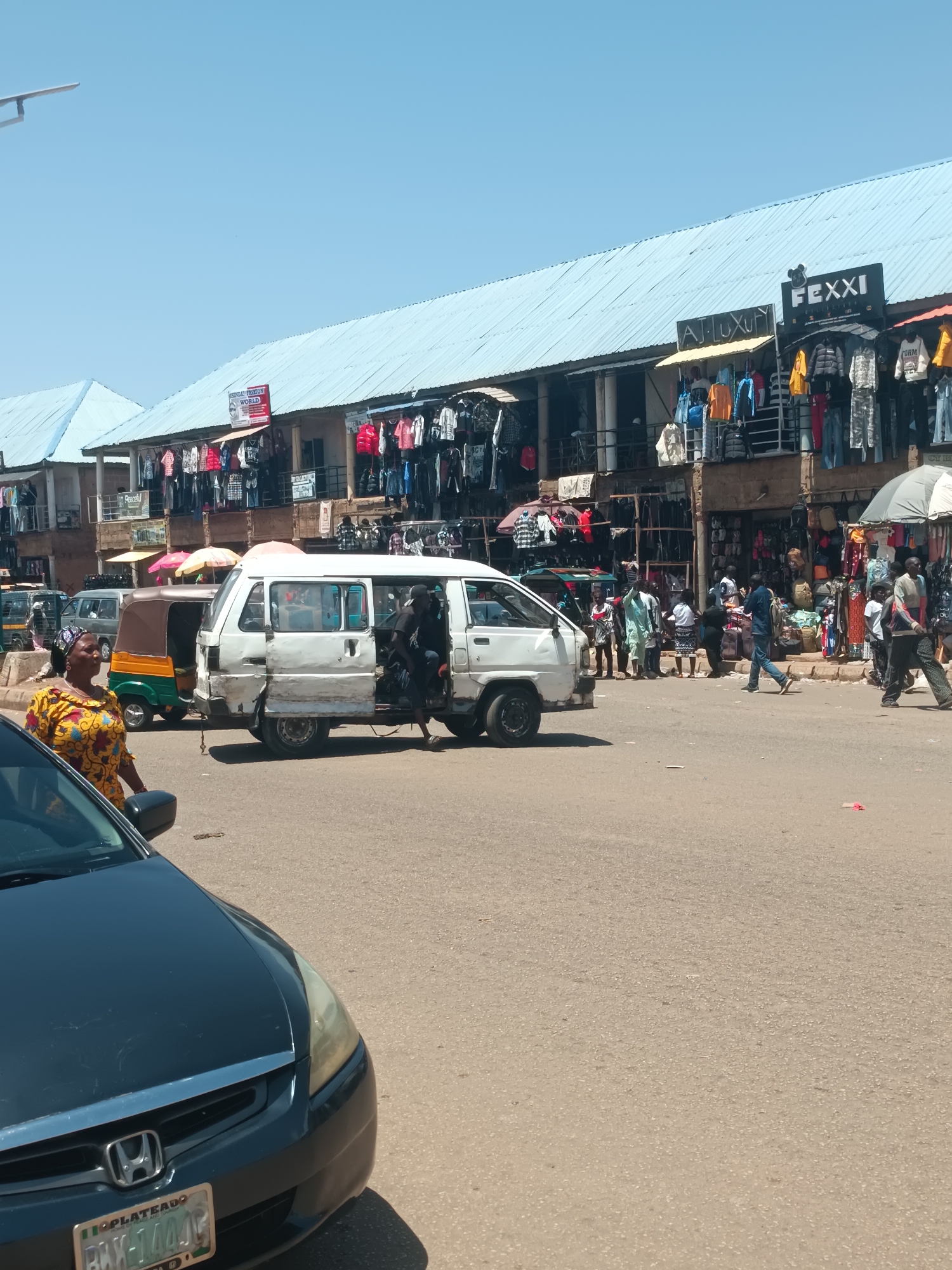 The image depicts a street scene in Bukuru, Nigeria, with a commercial building dominating the background. The building is two stories high, covered with a light blue corrugated metal roof, and has retail shops on the ground floor displaying various clothing items. Shop signs like "FEXXI" and "AJ LUXURY" are visible. People are milling around the storefronts, some browsing the merchandise.
In the foreground, a white passenger van with its door open is parked in the street. A person appears to be exiting the van. A black car, a Honda, is partially visible on the left side of the frame, with its Nigerian license plate reading "BXK-144AG" from Plateau State. A woman in a yellow and brown patterned top is visible near the front of the car. Other vehicles, including a motorcycle rickshaw, are also in the street. The road surface is paved. The sky is clear and blue.