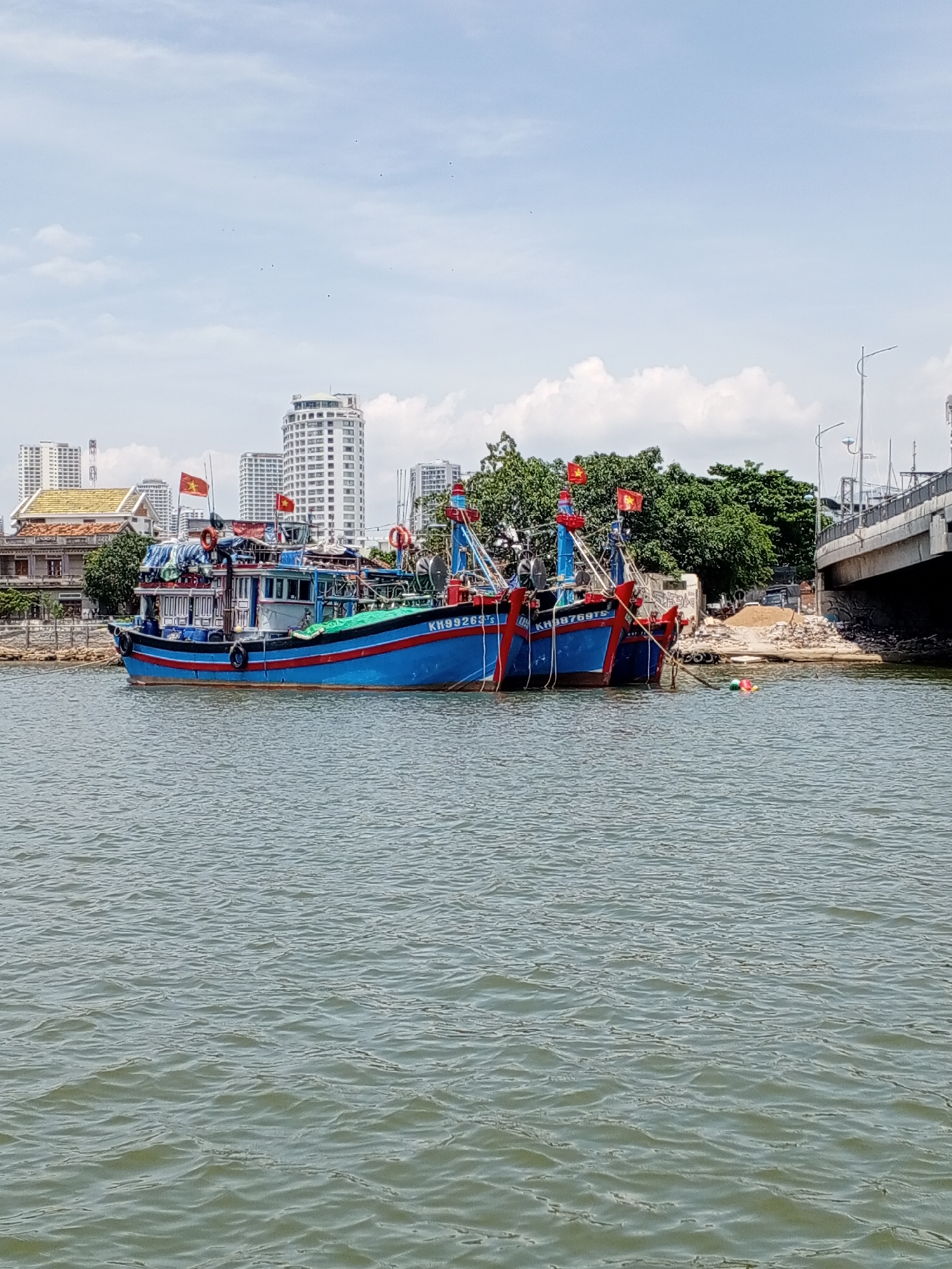 The image depicts a body of water with several blue fishing boats docked. The boats are adorned with red and blue stripes and have Vietnamese flags flying. In the background, a city skyline is visible with tall buildings and trees. A bridge crosses over the water in the right portion of the image. The sky is partly cloudy. The scene is likely located in Bac Nha Trang Ward, Vietnam, given the presence of Vietnamese flags and the architectural style of the buildings.