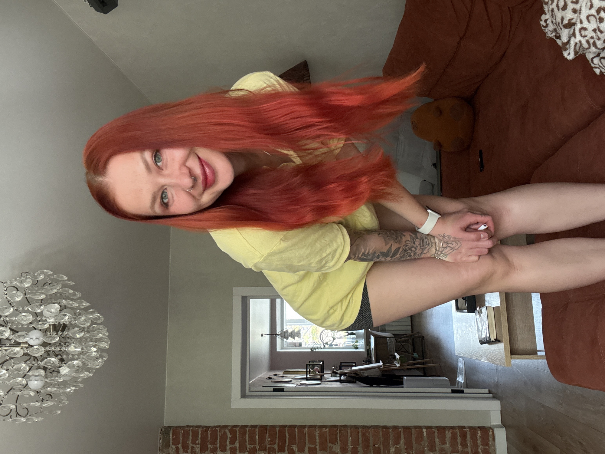 A woman with long, red hair is sitting on a brown couch. She is wearing a yellow t-shirt and dark shorts. She is smiling and looking at the camera. A crystal chandelier hangs from the ceiling. In the background, there is a hallway leading to a window. The location is potentially an apartment in Rostov-on-Don, Russia, based on the presence of brickwork, a common construction material in that area. The woman's arm has tattoos and she is wearing a white smartwatch. A coffee table is partially visible near the couch.