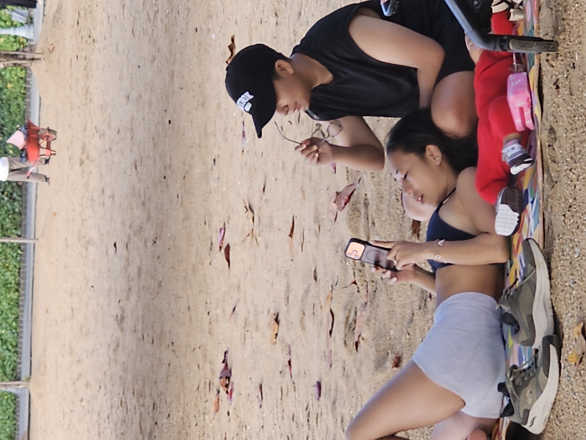 On a sandy beach, a man in a black tank top and black baseball cap examines a twig, while a woman in a black bikini top and light blue shorts uses a smartphone. Next to them, a toddler in red clothing lies on a colorful mat with various items. The location is likely a beach in Denpasar, Indonesia. The sand is strewn with small twigs and leaves. Vegetation and a metal fence are seen to the left.